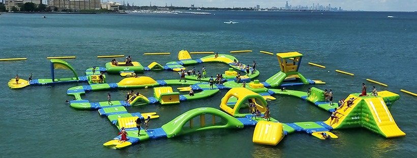 Large floating water park with various inflatable slides, climbing structures, and obstacle courses on the water, with children and adults playing.