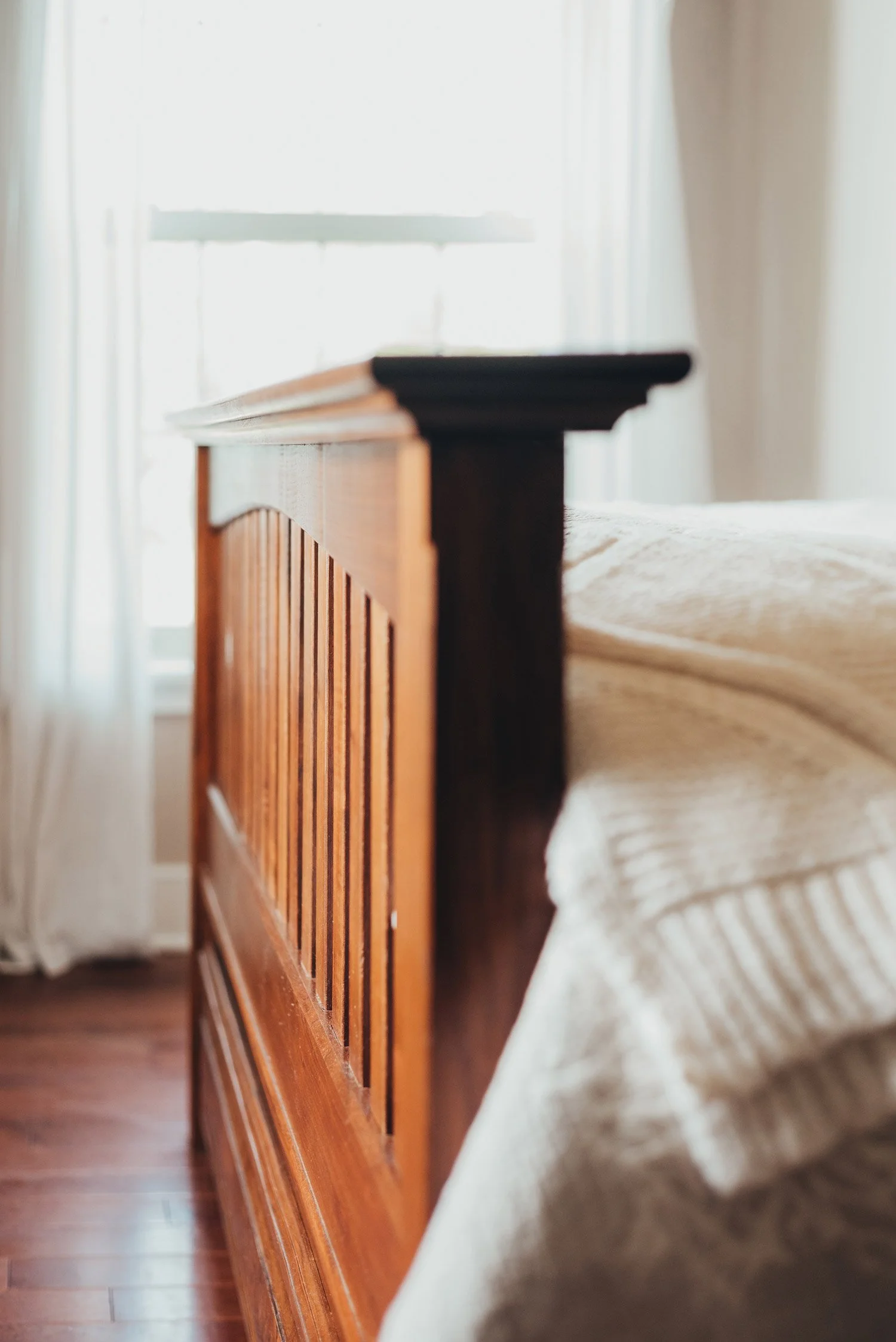 Close-up of a wooden bed frame next to a beige bedspread, with a window in the background.