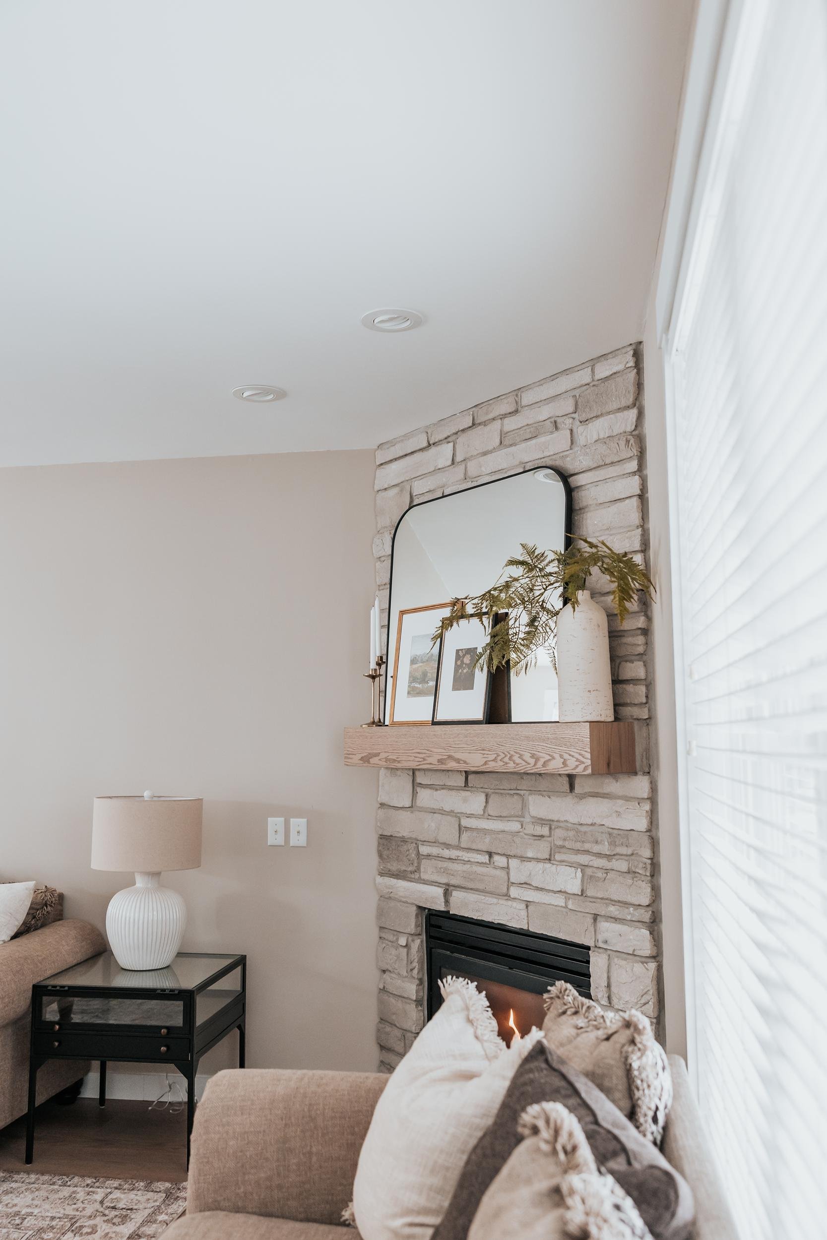 Living room with beige sofas, black side table with a white lamp, brick fireplace with a wooden mantel, mirror, framed photos, and decorative vases with greenery.