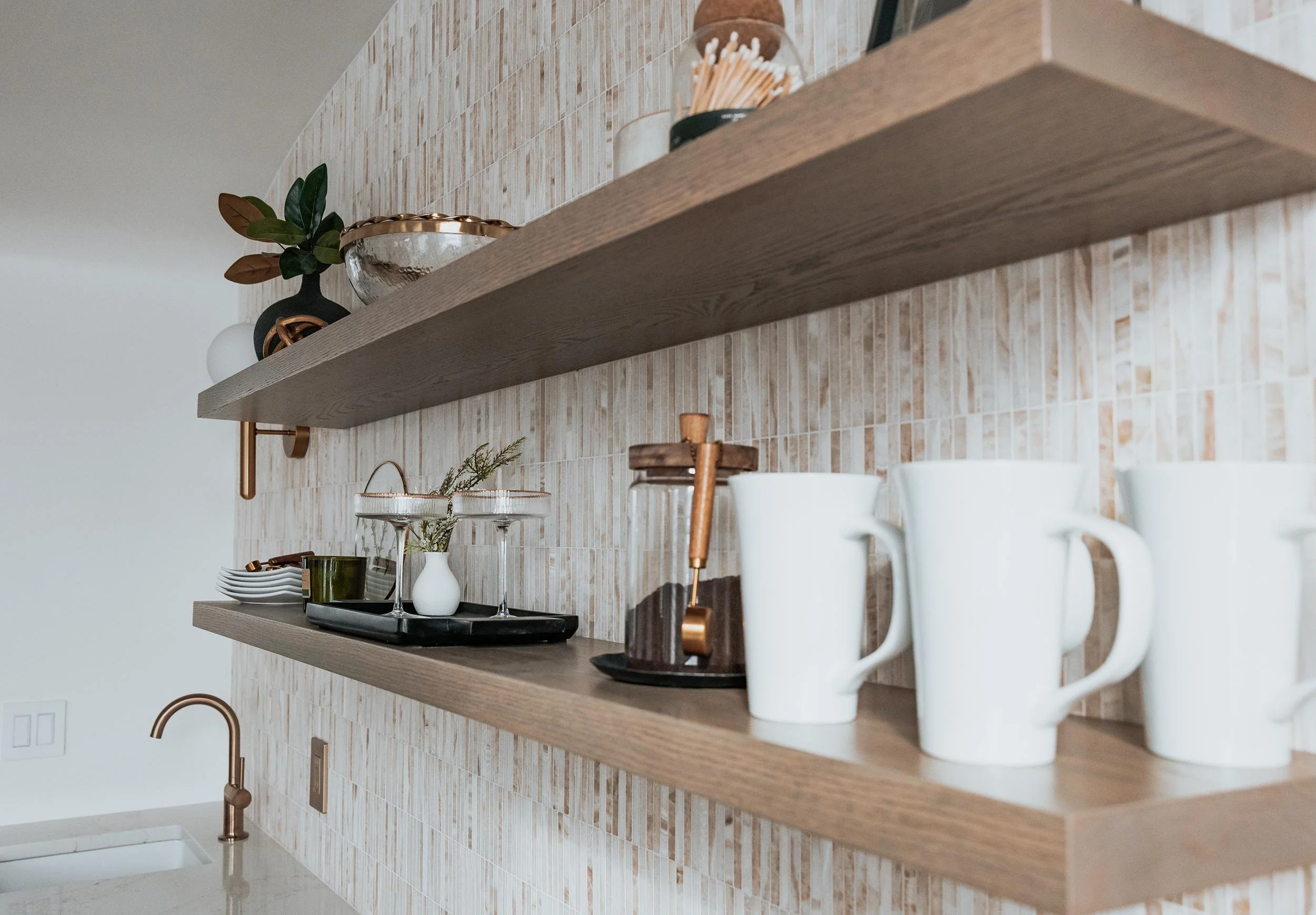 Open wooden kitchen shelves holding glassware, mugs, a small plant, and decorative items in front of a tiled backsplash.
