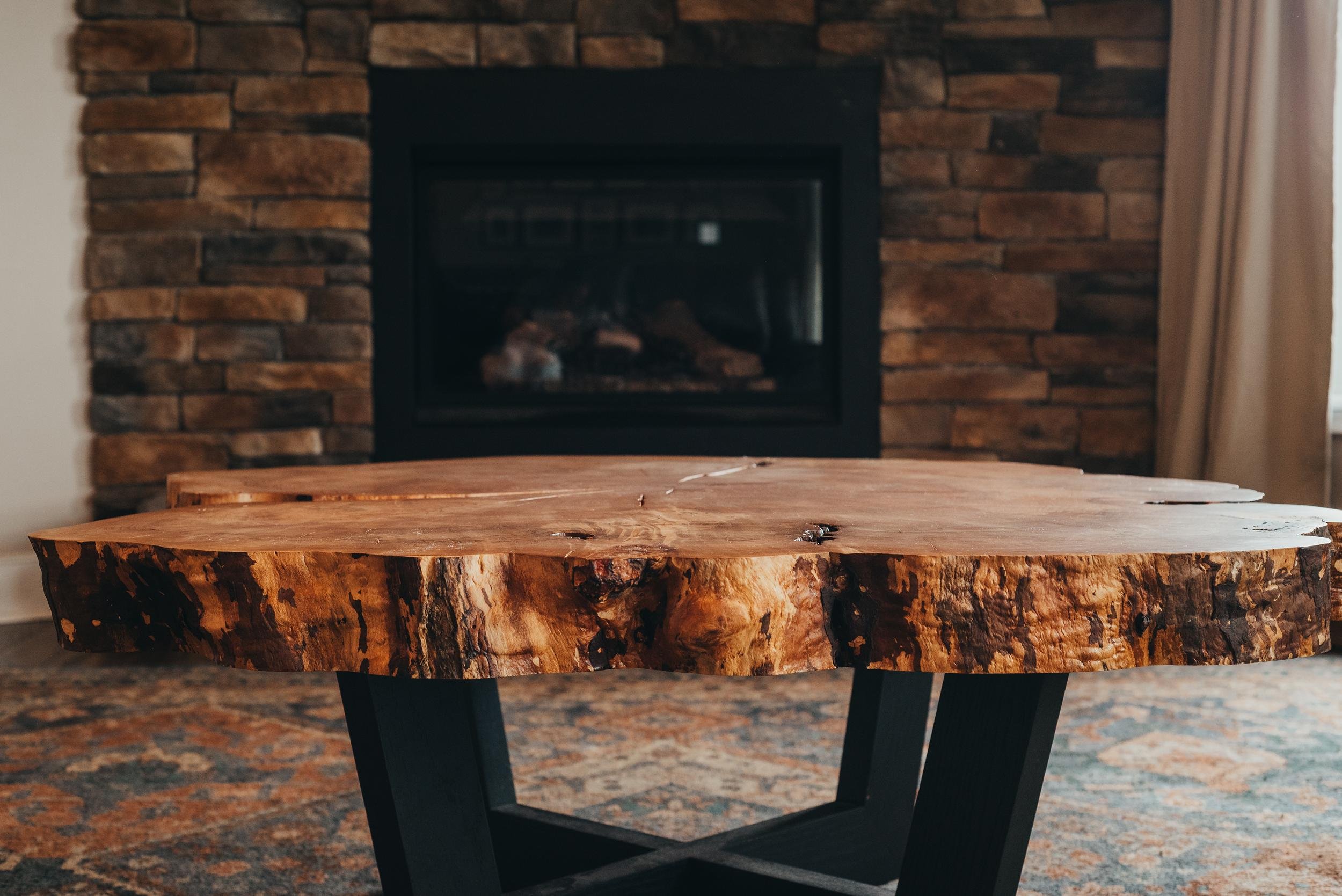 A rustic wooden coffee table with a natural edge, sitting on a patterned carpet in front of a brick fireplace with a black frame. Light coming through a curtain on the right.