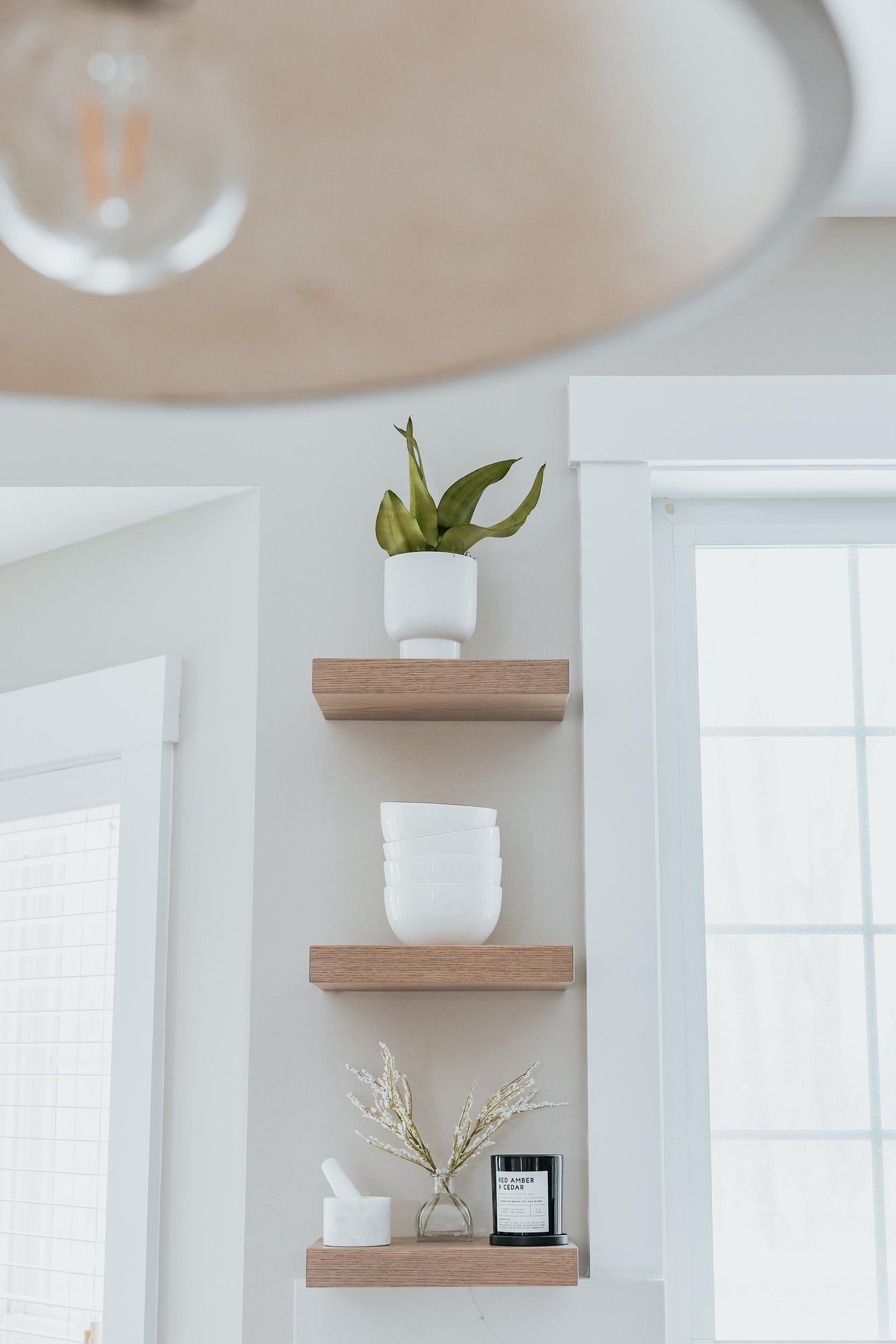 White wall with three floating wooden shelves holding a potted plant, white bowls, a mortar and pestle, a candle, and a glass vase with dried flowers, next to a window with white trim.