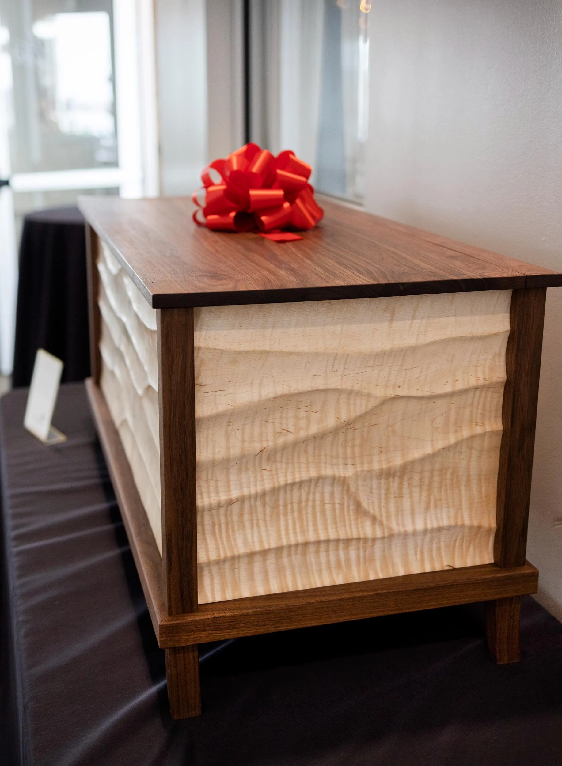 Wooden chest with wavy carved panels on the sides, topped with a red decorative bow.