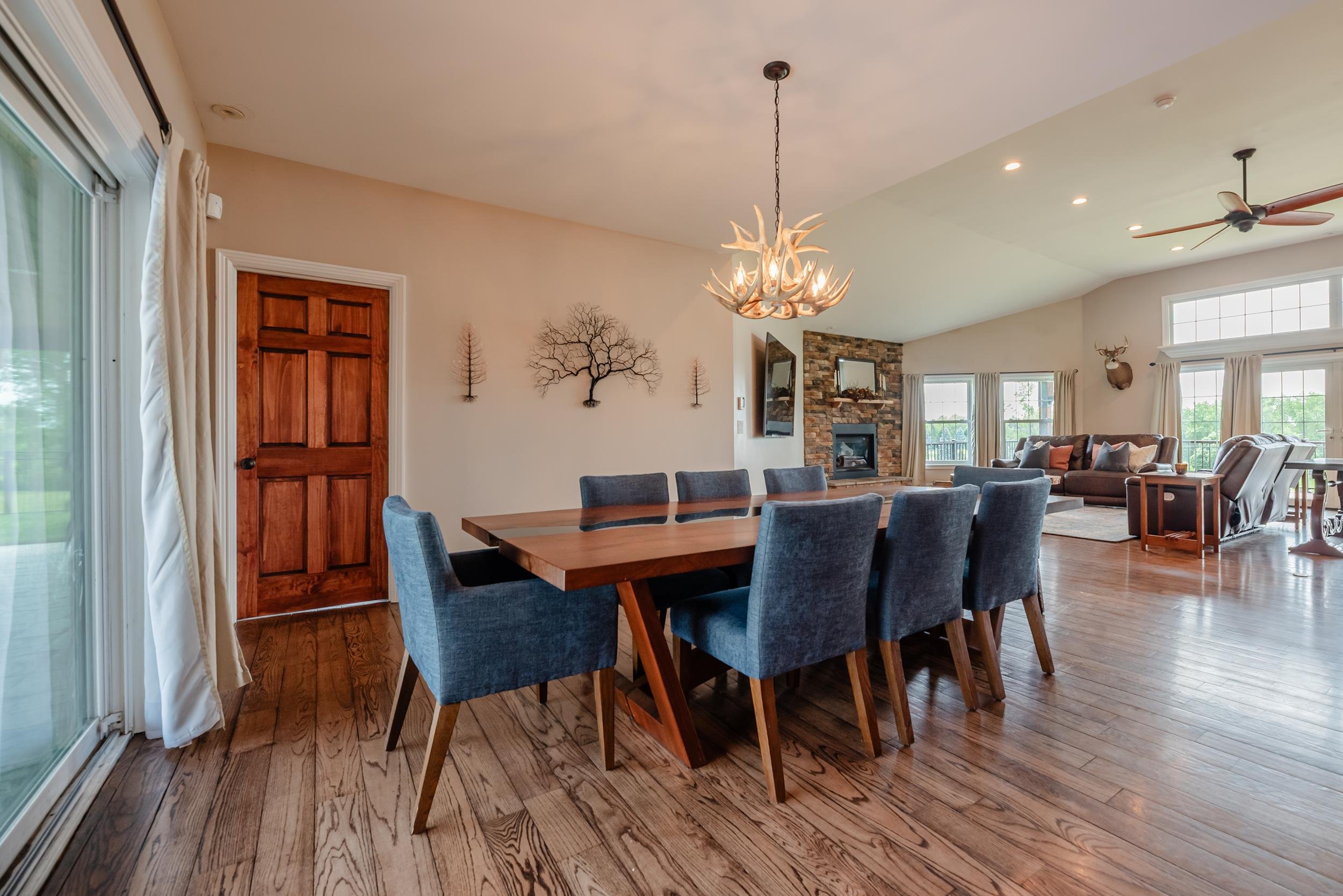 Open-concept living and dining room with wooden flooring, a large wooden dining table with blue upholstered chairs, a stone fireplace, large windows with curtains, a ceiling fan, and wall decor including a deer head and wall-mounted sculptures.