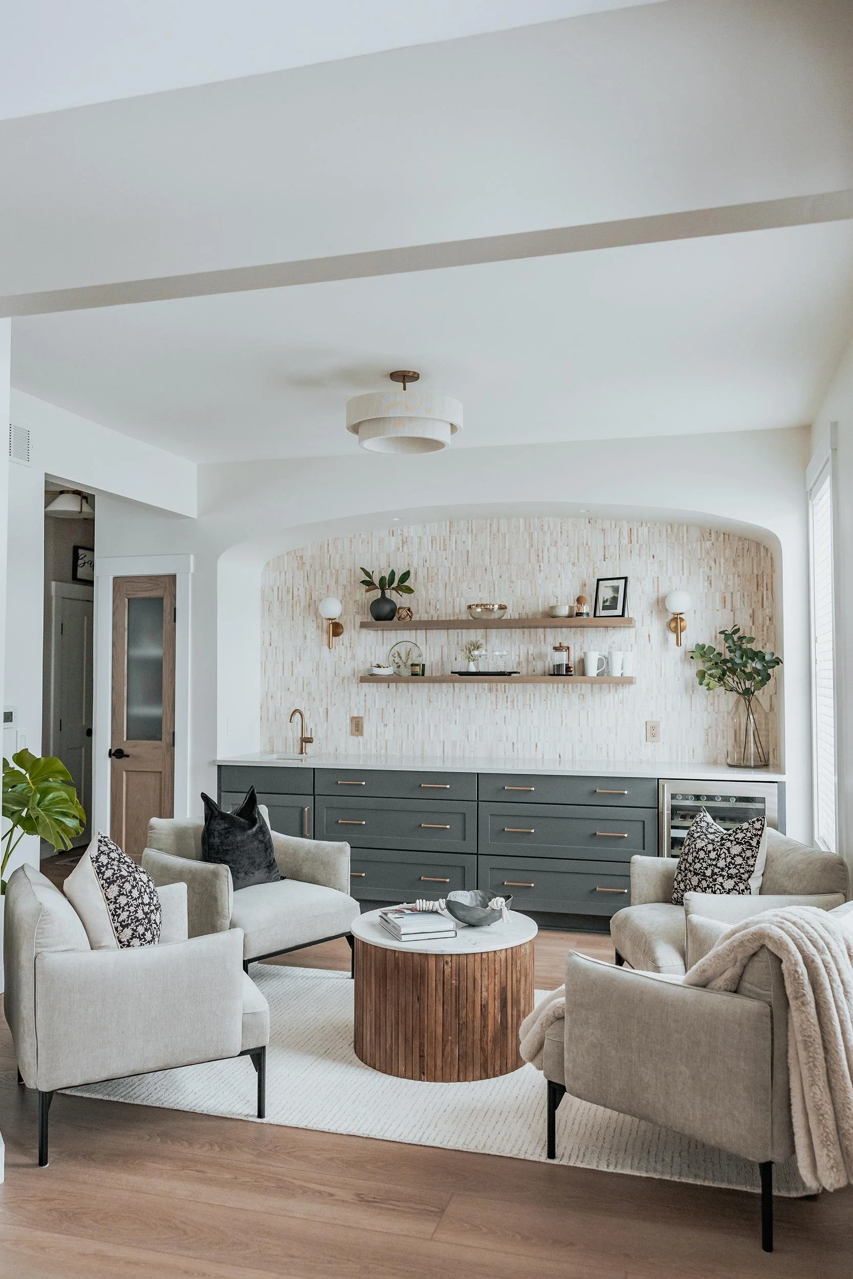 Living room with beige sofas, a round wooden coffee table, gray cabinets with gold handles, open shelves with decorative items, and a light-colored textured wall.