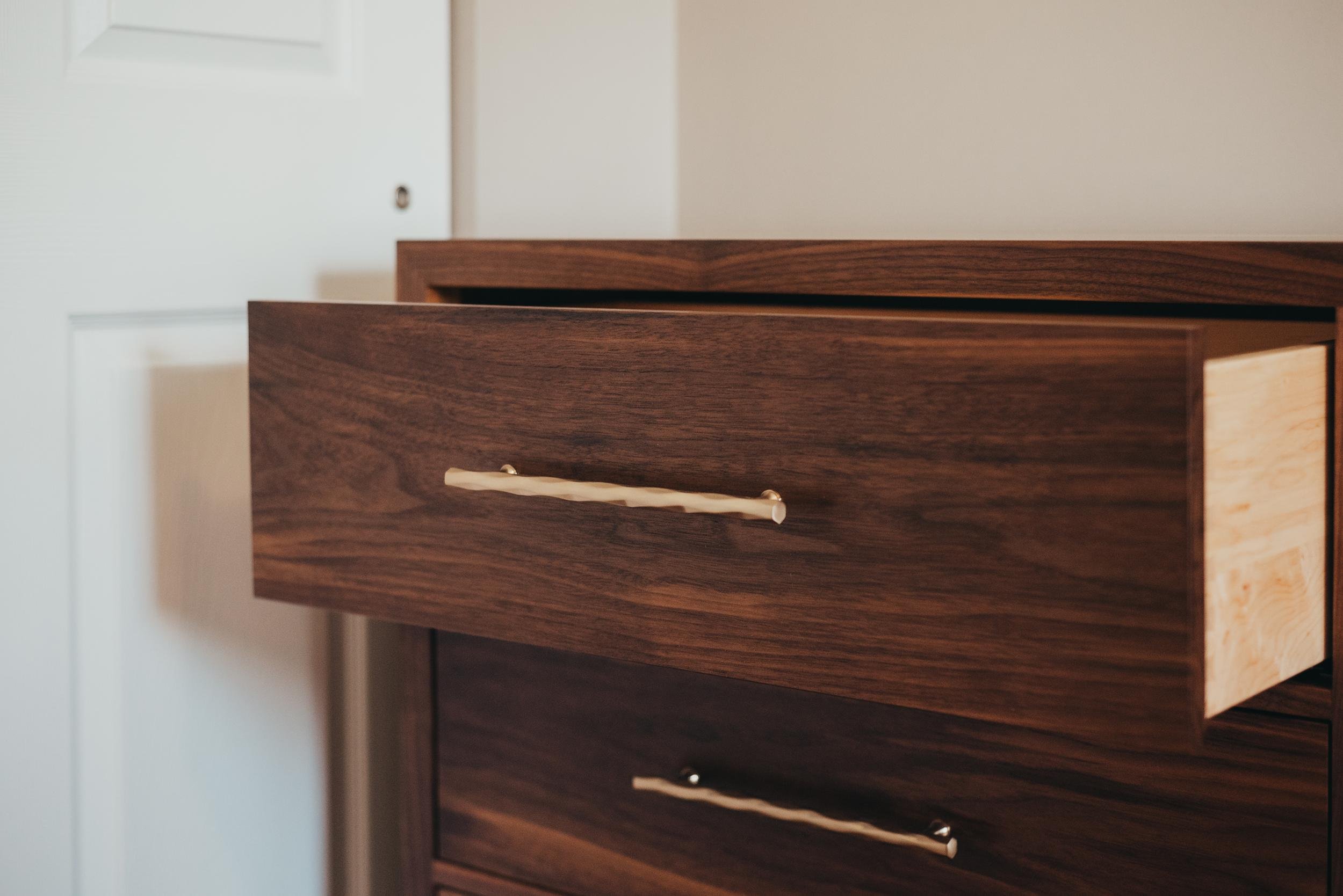 Close-up of a wooden dresser drawer with woven pattern handles in a room with white door and beige wall.