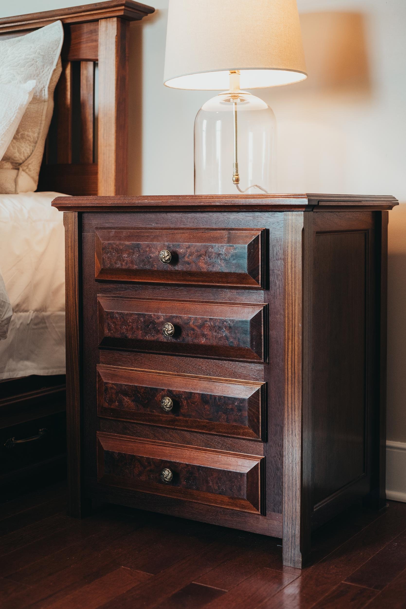 A wooden bedside table with four drawers, a glass table lamp on top, next to a bed with pillows and a wooden headboard, on a hardwood floor.