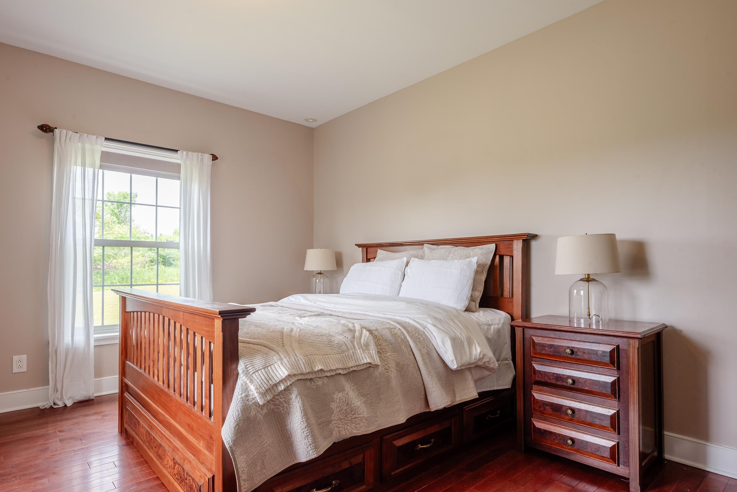 A neatly made bed with white bedding and pillows, a wooden headboard, and matching wooden nightstands with lamps in a bedroom with beige walls and a window with white curtains.