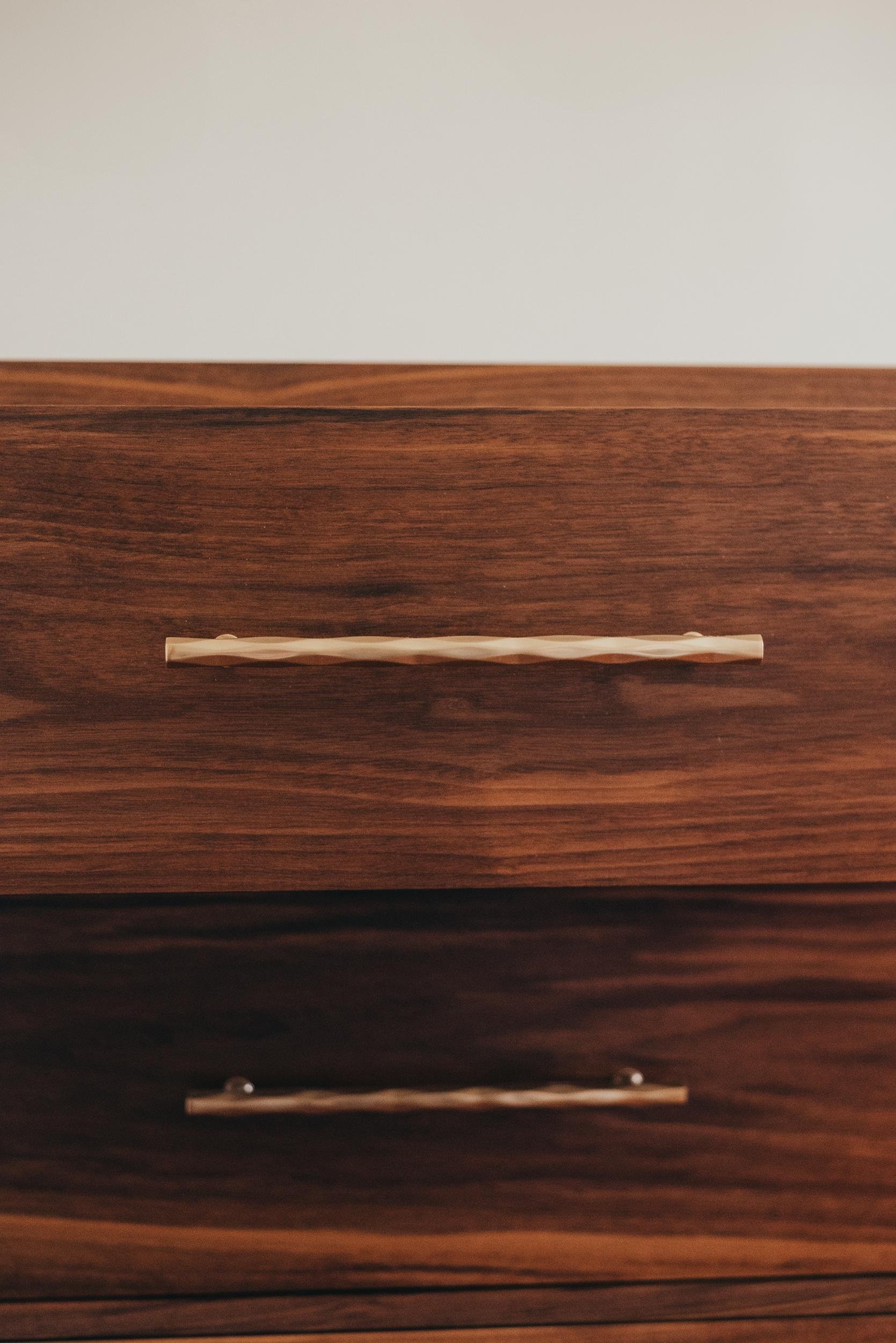 Close-up of a wooden drawer with a horizontal, twisted metal handle, against a plain, light-colored wall.