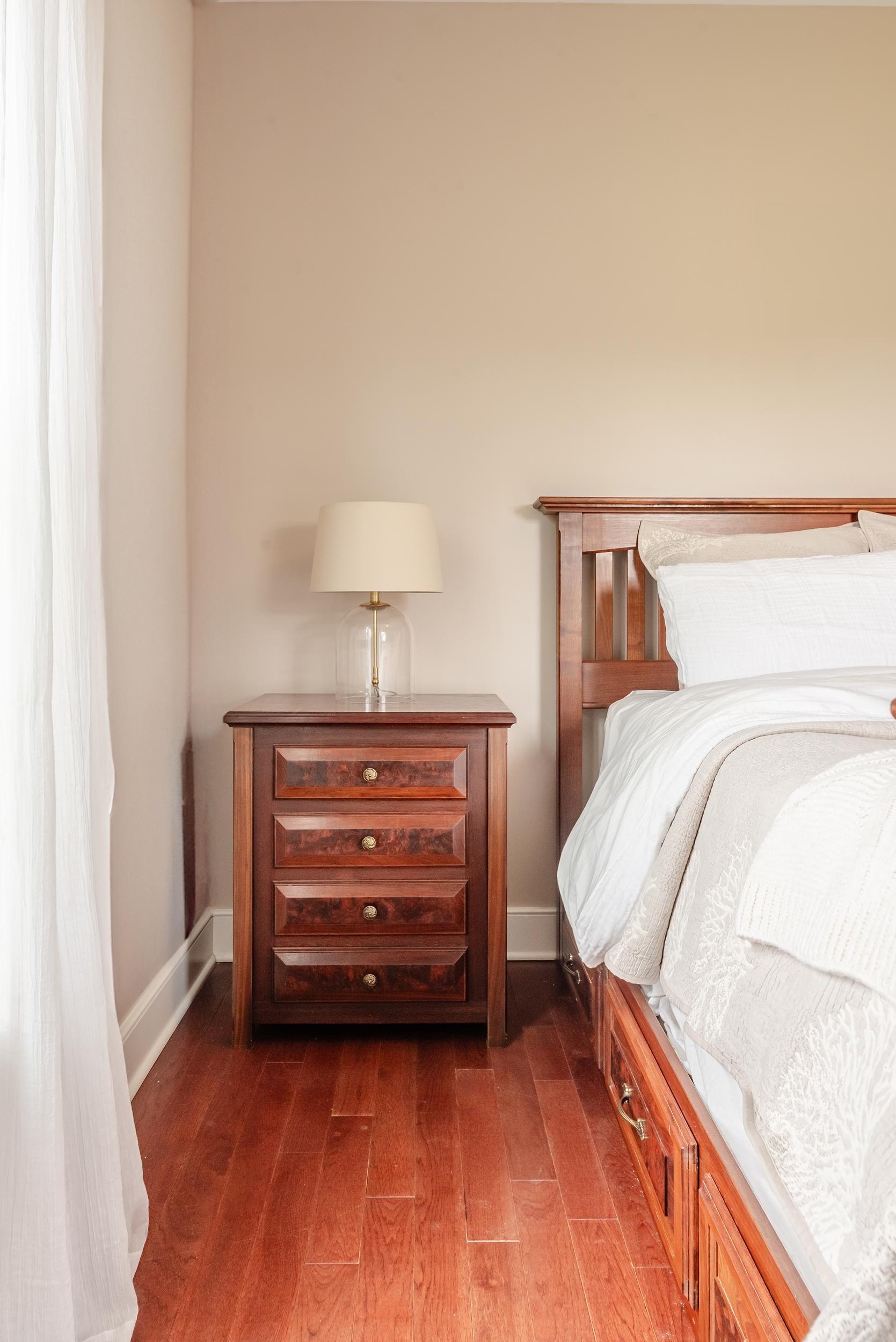 A bedroom scene with a wooden nightstand with four drawers and a brass knob, a glass table lamp with a white shade, a bed with a wooden headboard, white bedding, and hardwood flooring.