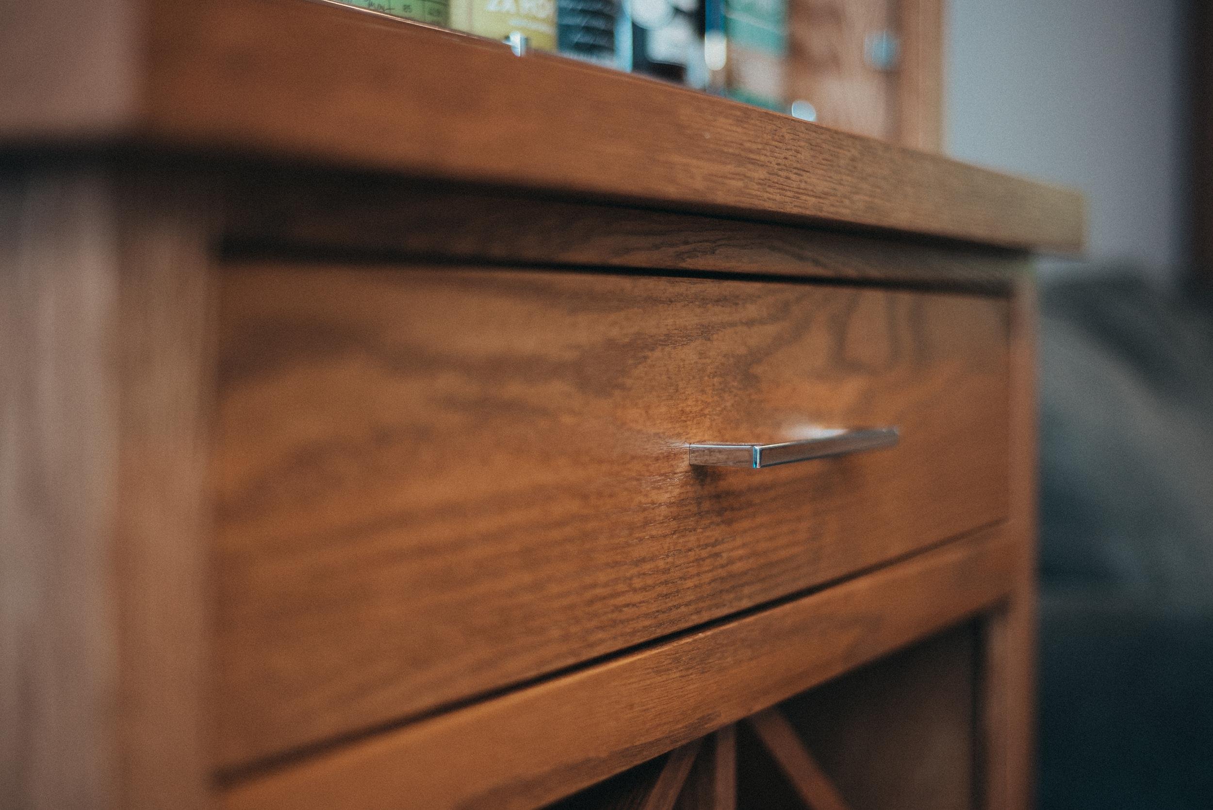 Close-up of a wooden dresser drawer with a metallic handle, showing wood grain and part of the top surface with some books or objects on it.