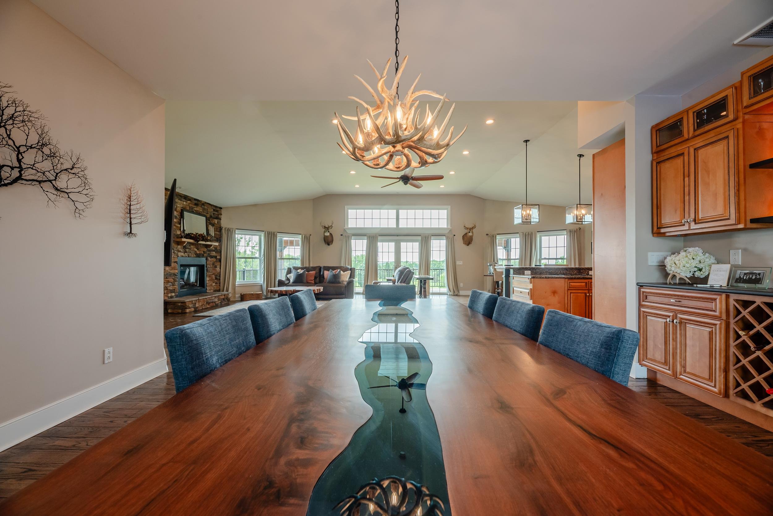 Interior view of a spacious dining and living area with a long wooden table and blue chairs, a chandelier made of antlers, a fireplace with a stone surround, large windows with curtains, and mounted deer heads on the walls.