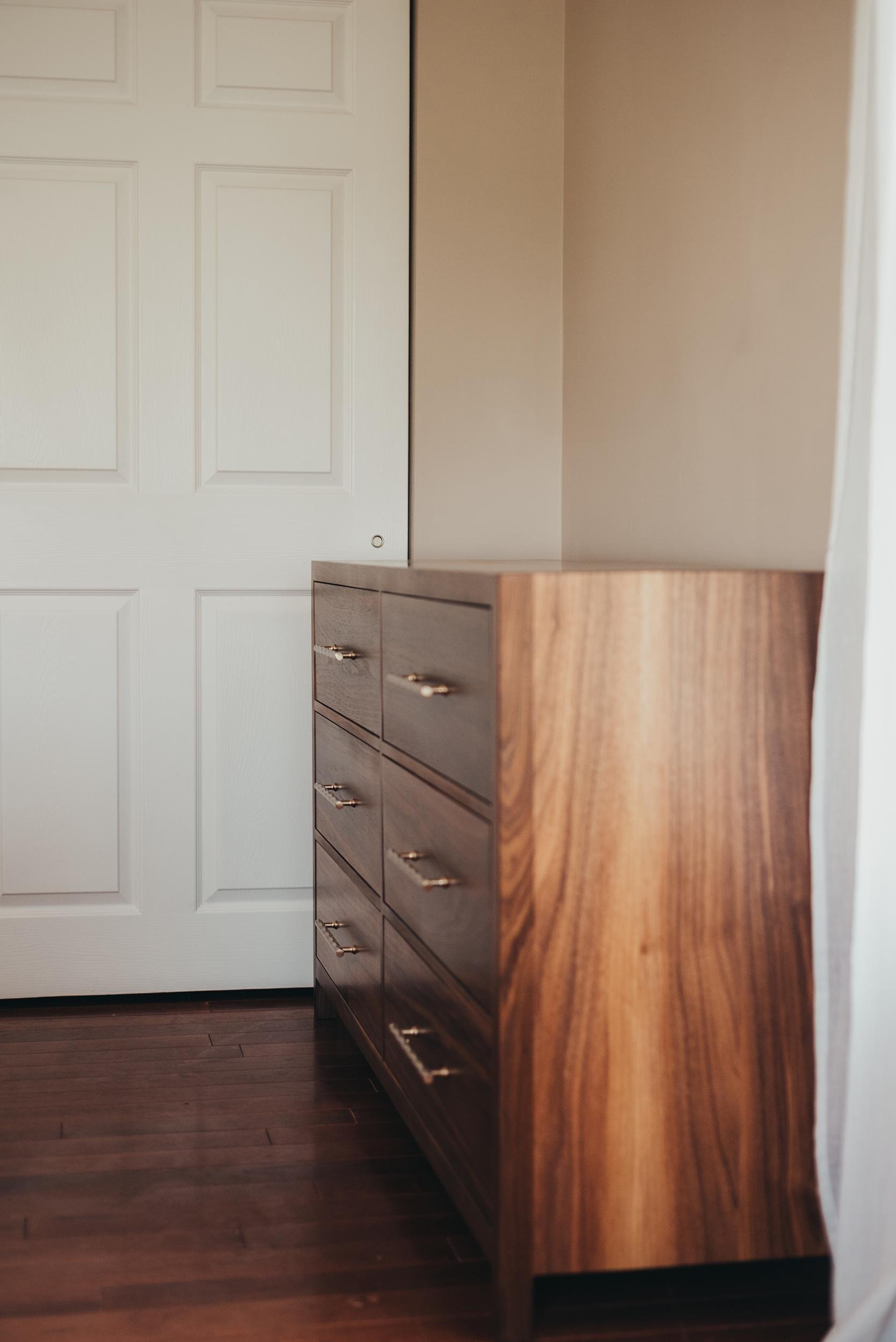 A wooden dresser with multiple drawers and gold handles, positioned next to a white closet door in a room with beige walls and dark hardwood floors.