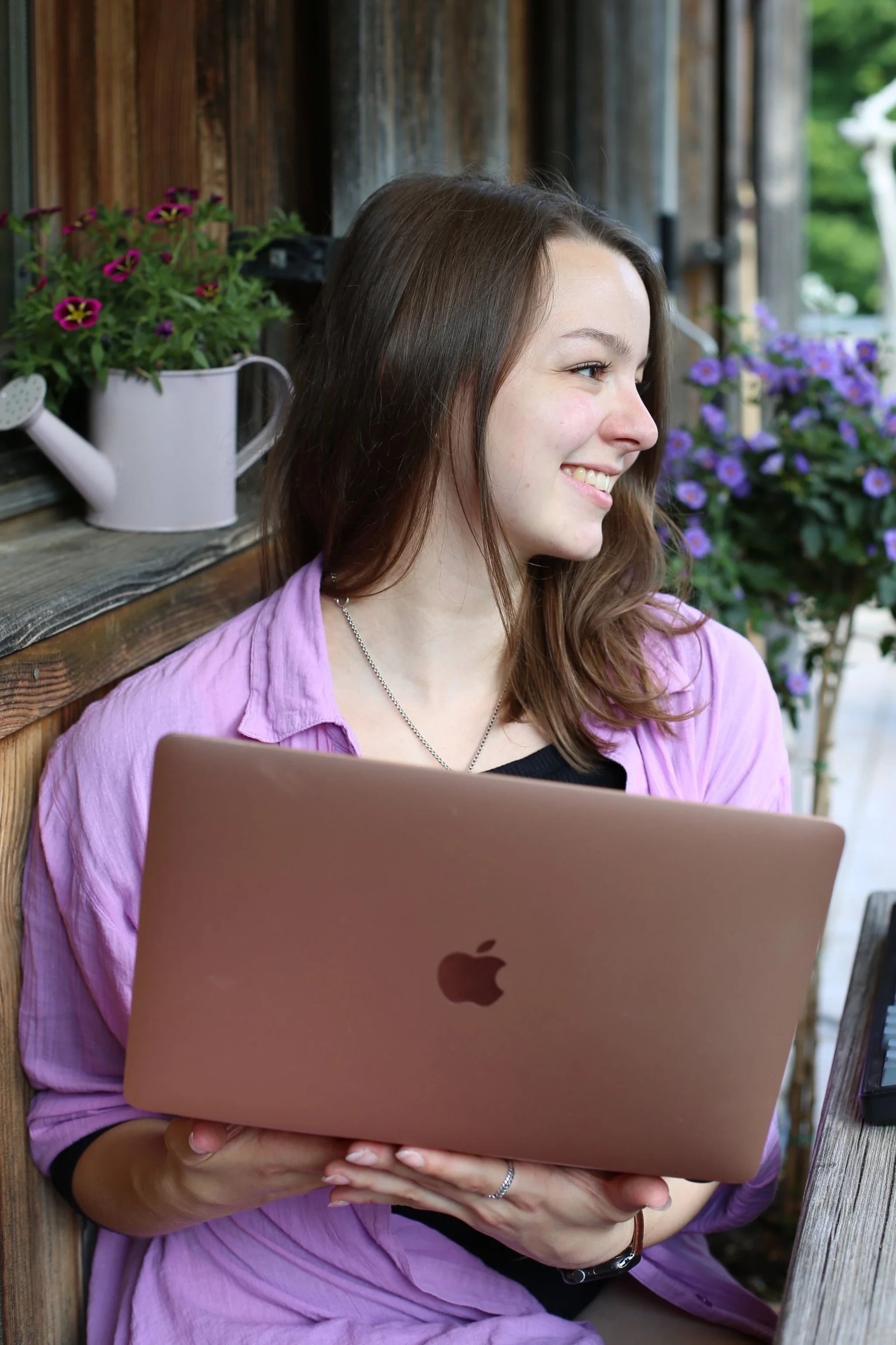 Junge Frau mit braunen Haaren, trägt eine rosa Jacke, hält einen rosegoldfarbenen MacBook und sitzt im Freien neben Blumen und einer Holzwand.