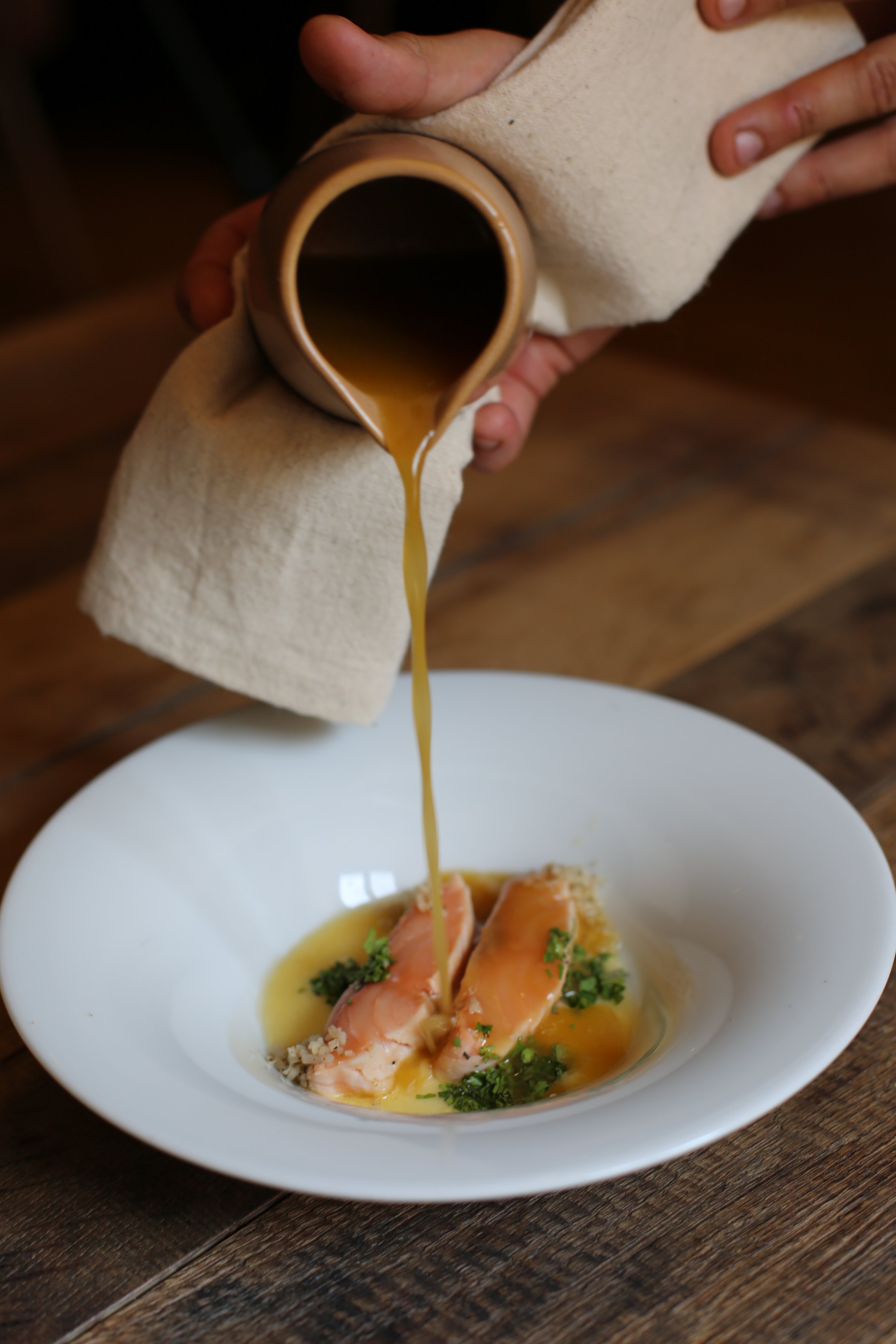 Jemand gießt Sauce aus einer Kanne auf Lachs mit Kräutern auf einem weißen Teller.