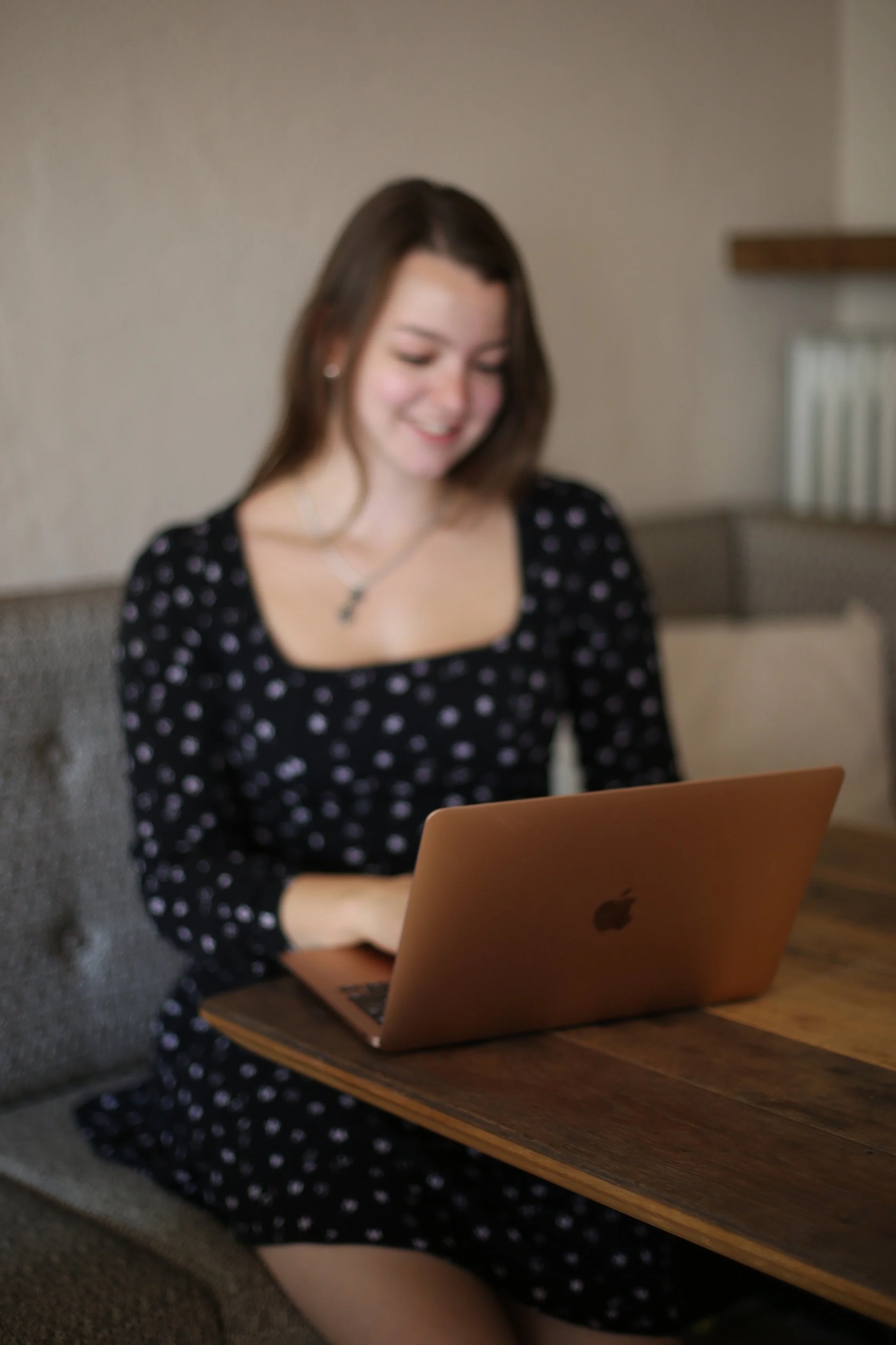 Eine junge Frau sitzt an einem Holztisch und arbeitet mit einem rosegoldfarbenen Laptop in einem gemütlichen Raum.
