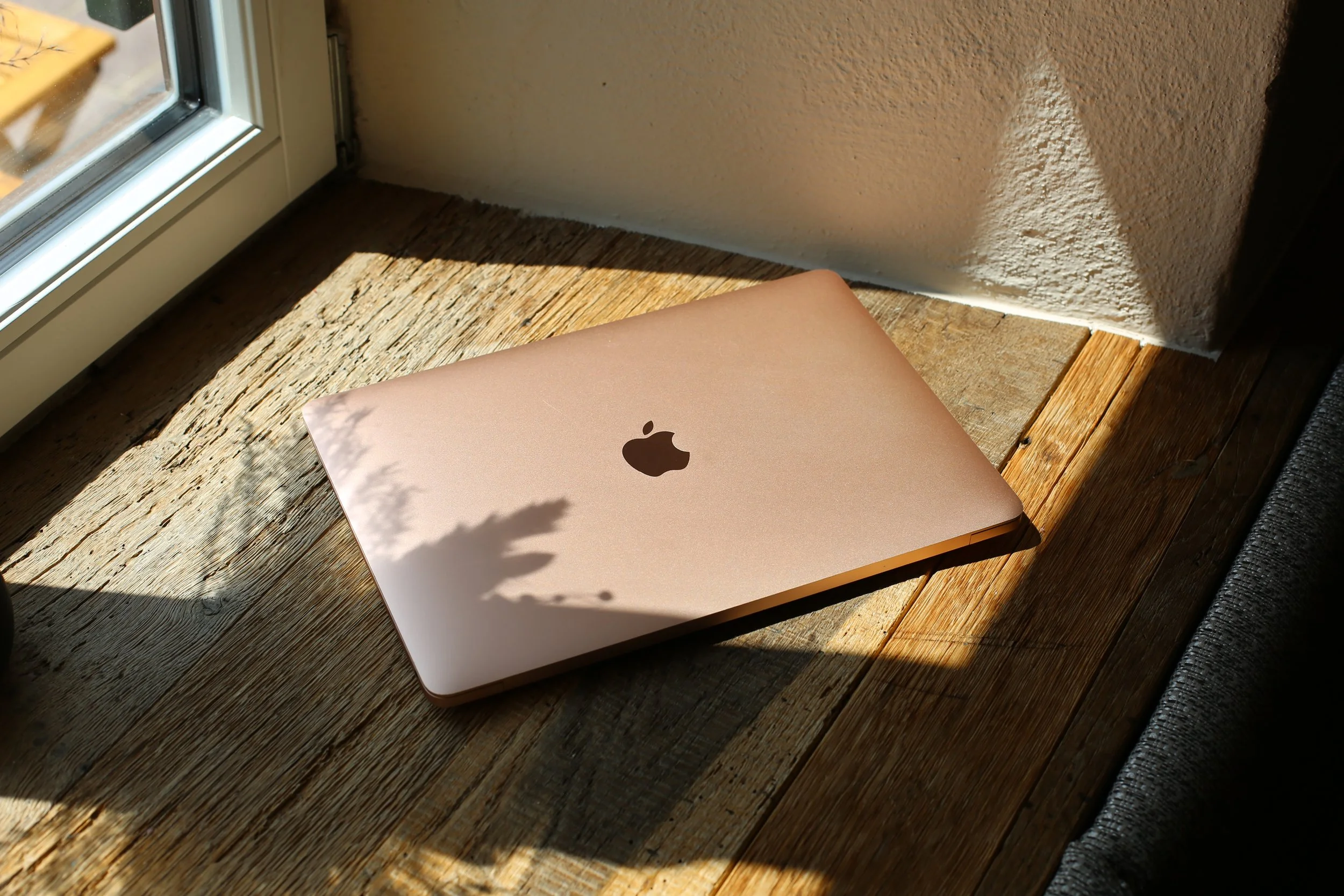 Ein rosegoldfarbener MacBook Laptop liegt auf einer Holzfensterbank im Sonnenlicht neben einer weißen Wand.