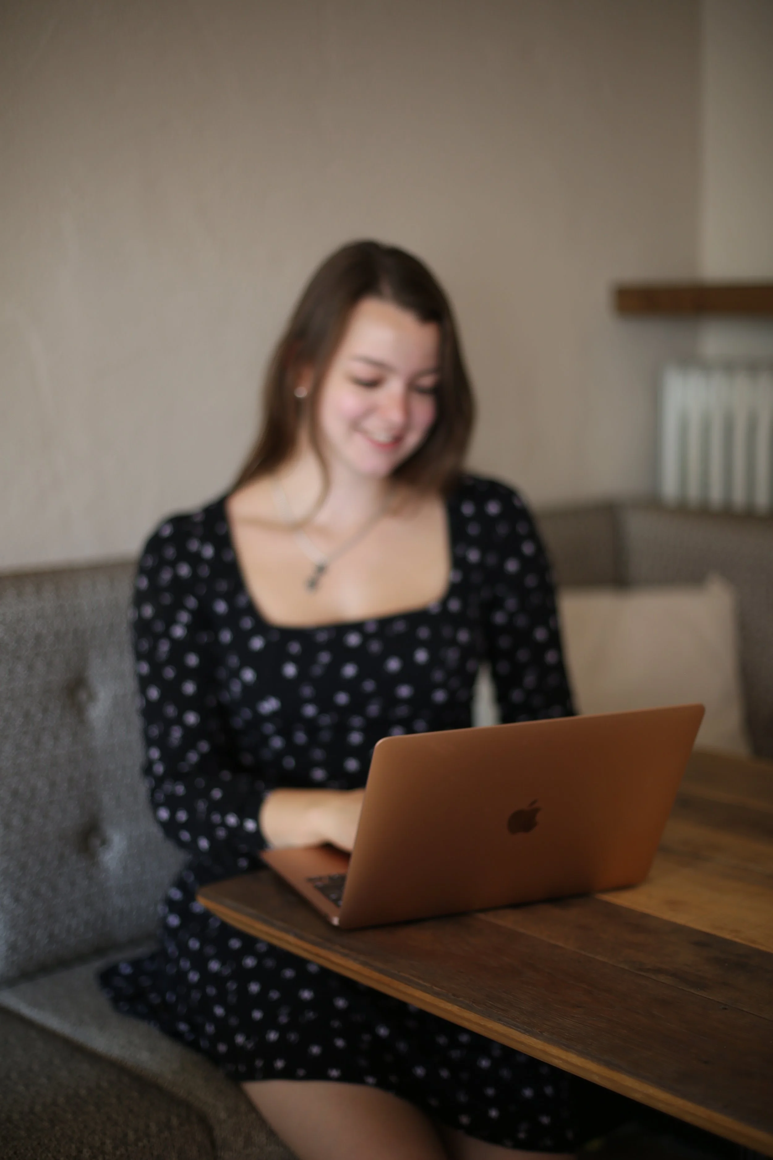 Junge Frau sitzt in einem gemütlichen Raum an einem Holztisch, arbeitet an einem rosegoldfarbenen MacBook, trägt einen schwarzen Kleid mit weißen Punkten und hat lange braune Haare.