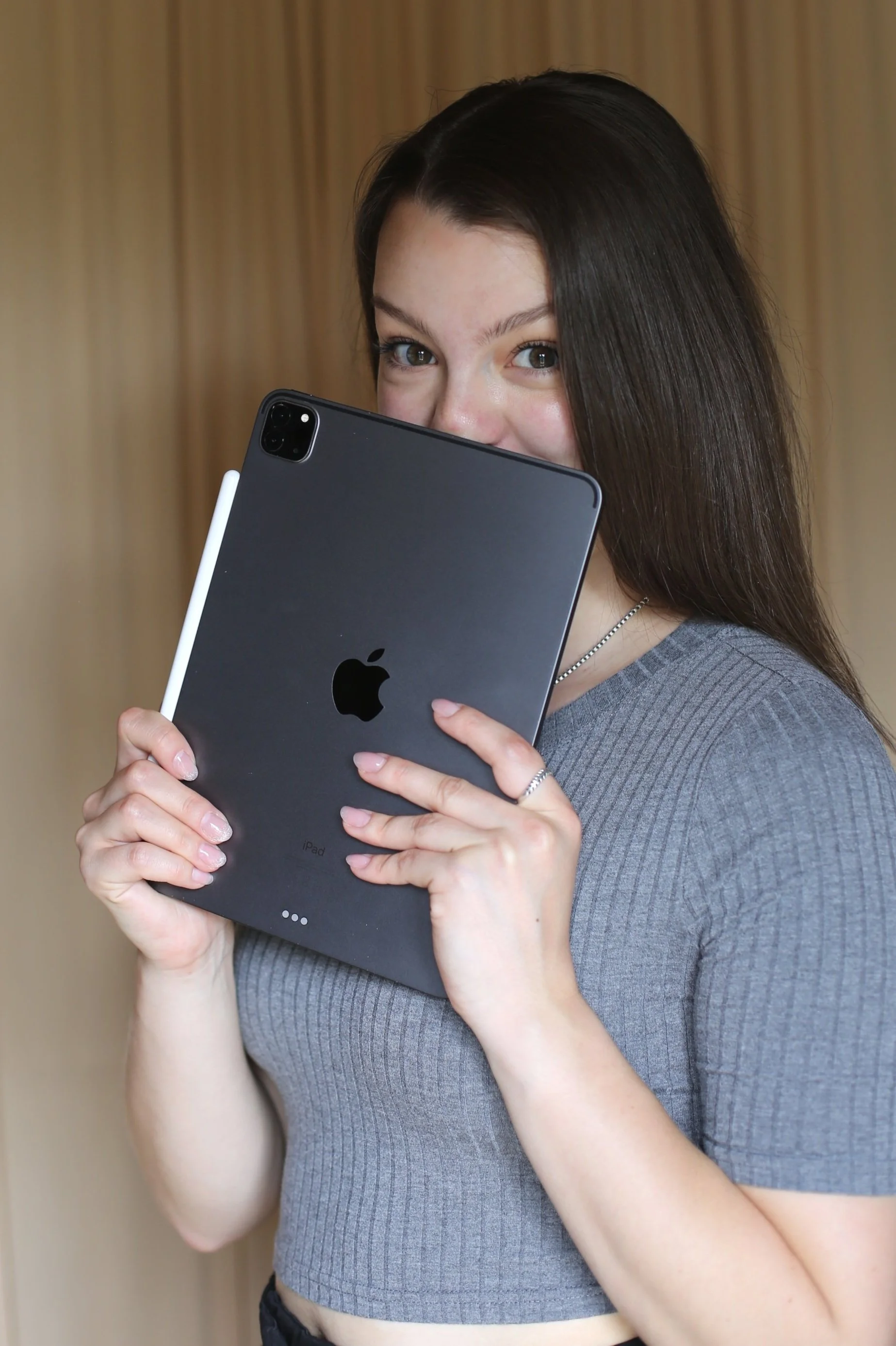 Junge Frau hält ein Apple iPad und versteckt ihr Gesicht hinter dem Gerät. Sie trägt ein graues Oberteil und hat braune Haare.