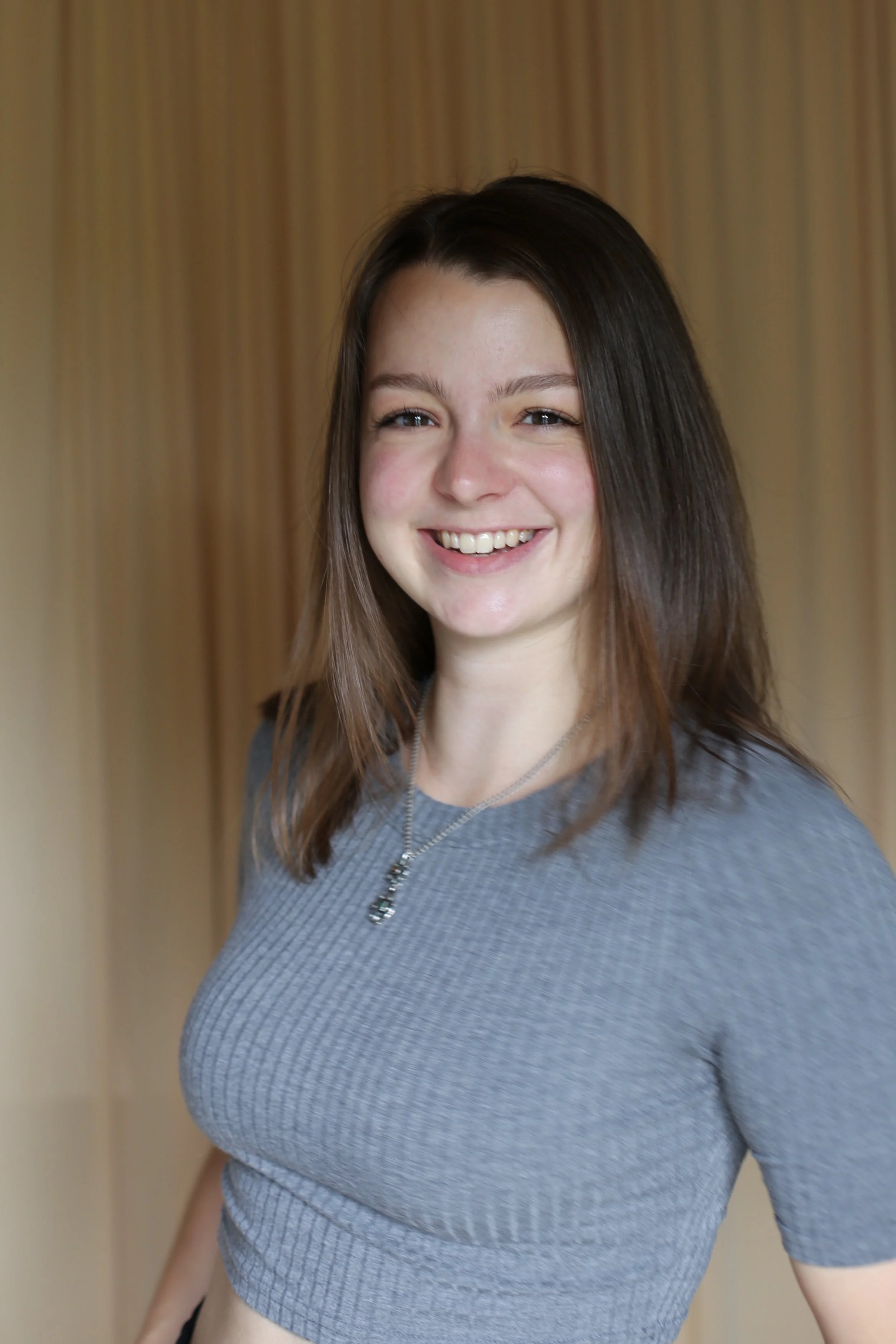 Junge Frau mit langem braunem Haar, graues T-Shirt und Kette, lächelnd, im Hintergrund eine holzvertäfelte Wand.