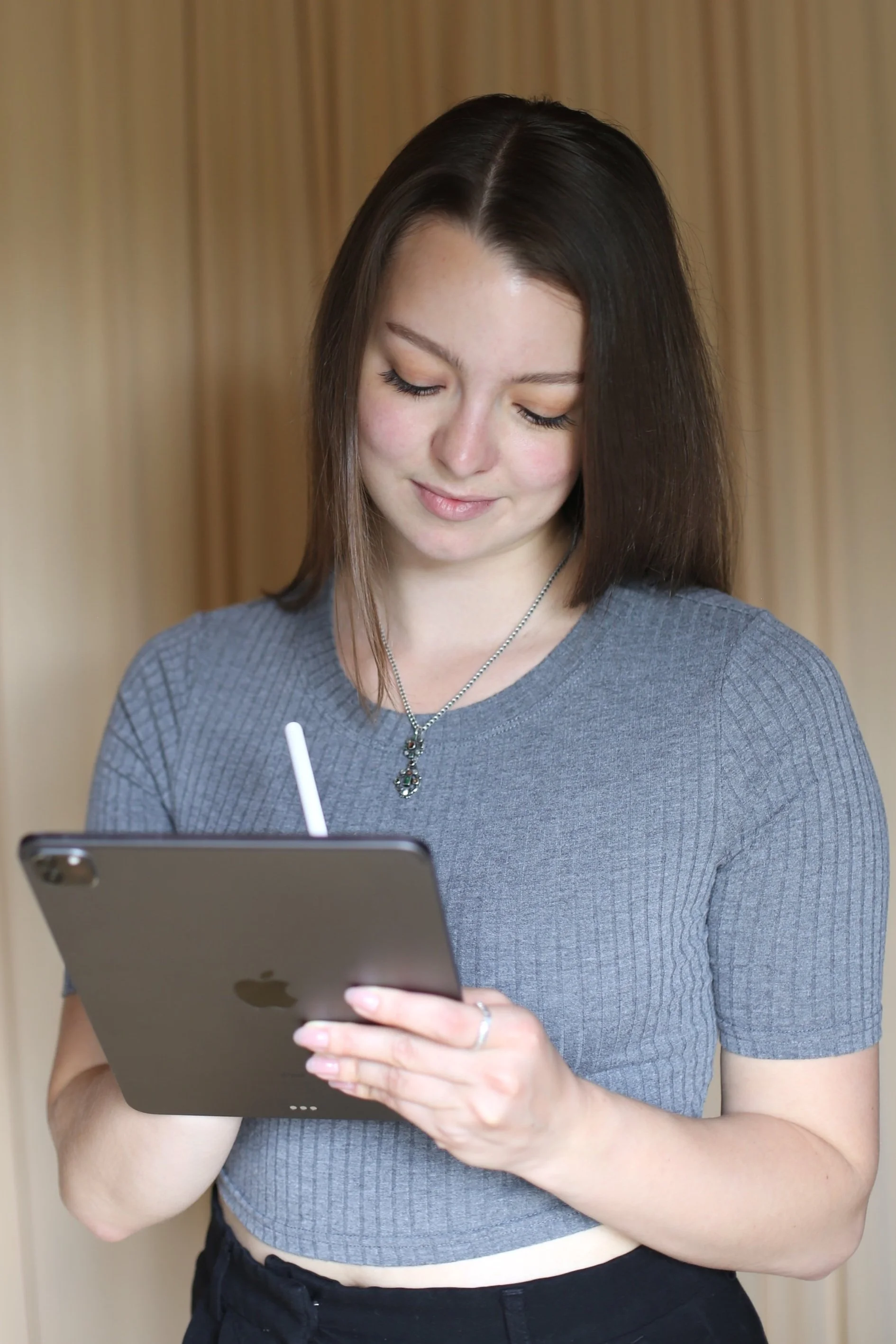 Junge Frau mit dunklem Haar, grauem T-Shirt und Kette, die auf einem iPad schreibt.