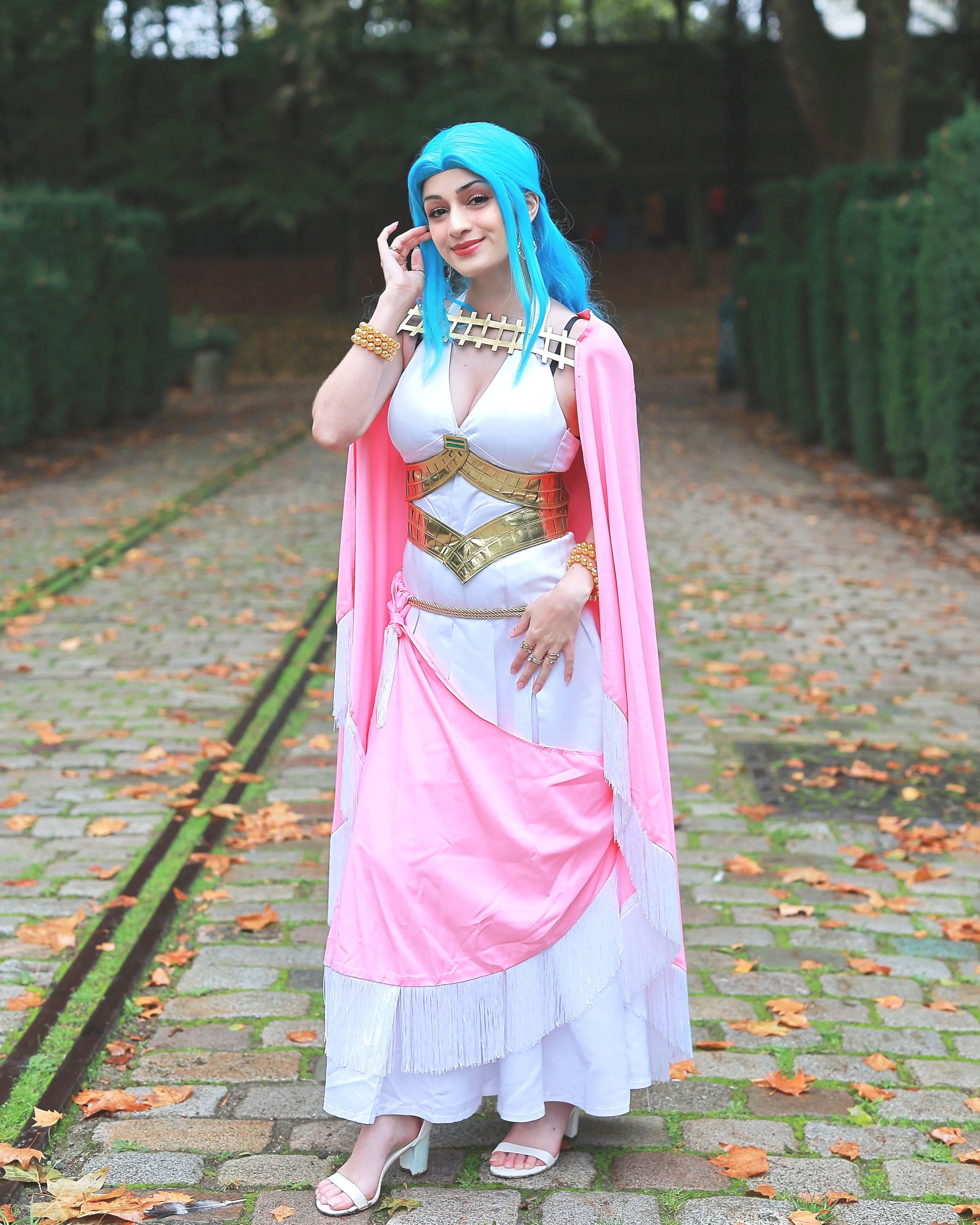 A woman in a colorful costume with blue hair and a pink and gold cape standing on a cobblestone path surrounded by green trees. cosplay cosplayer anime manga