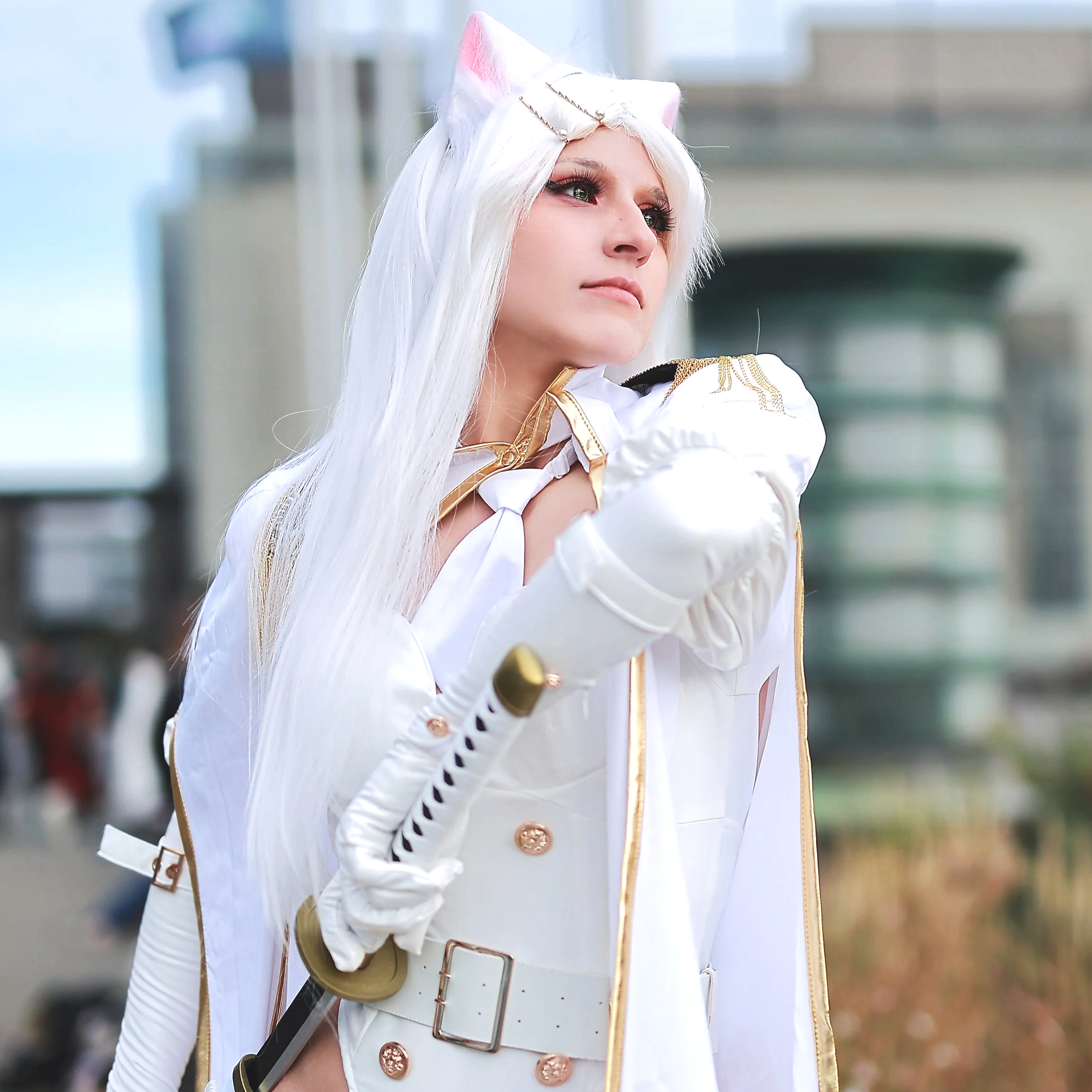 Person dressed as a fantasy character with long white hair, cat ears headpiece, white and gold costume, holding a sword, outdoors in a city environment. Cosplay anime manga gaming