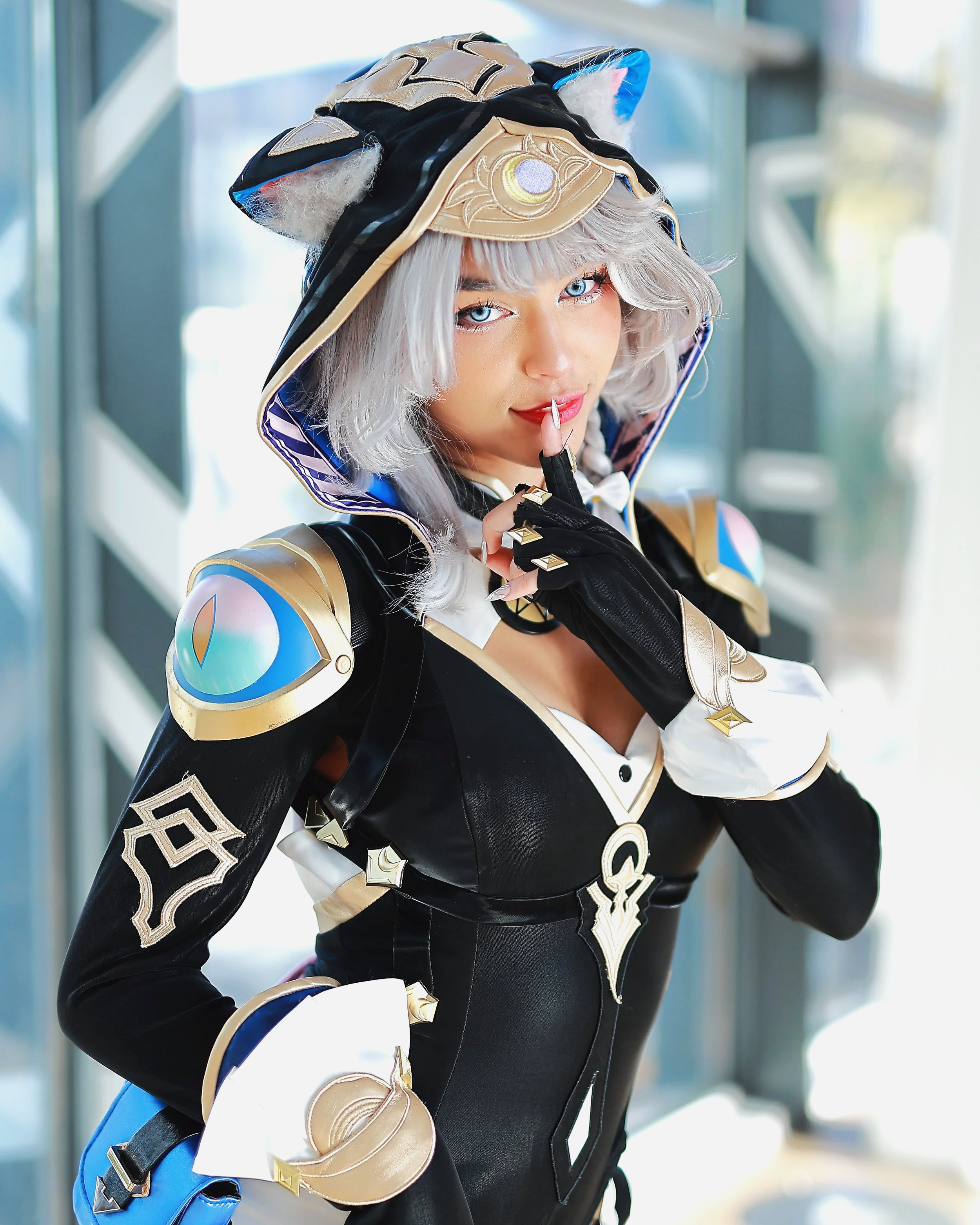 Une femme cosplaying un personnage avec une tenue noire et or, un capuchon avec des oreilles de chat, et des accessoires en forme d'œil, faisant un signe de silence avec le doigt.