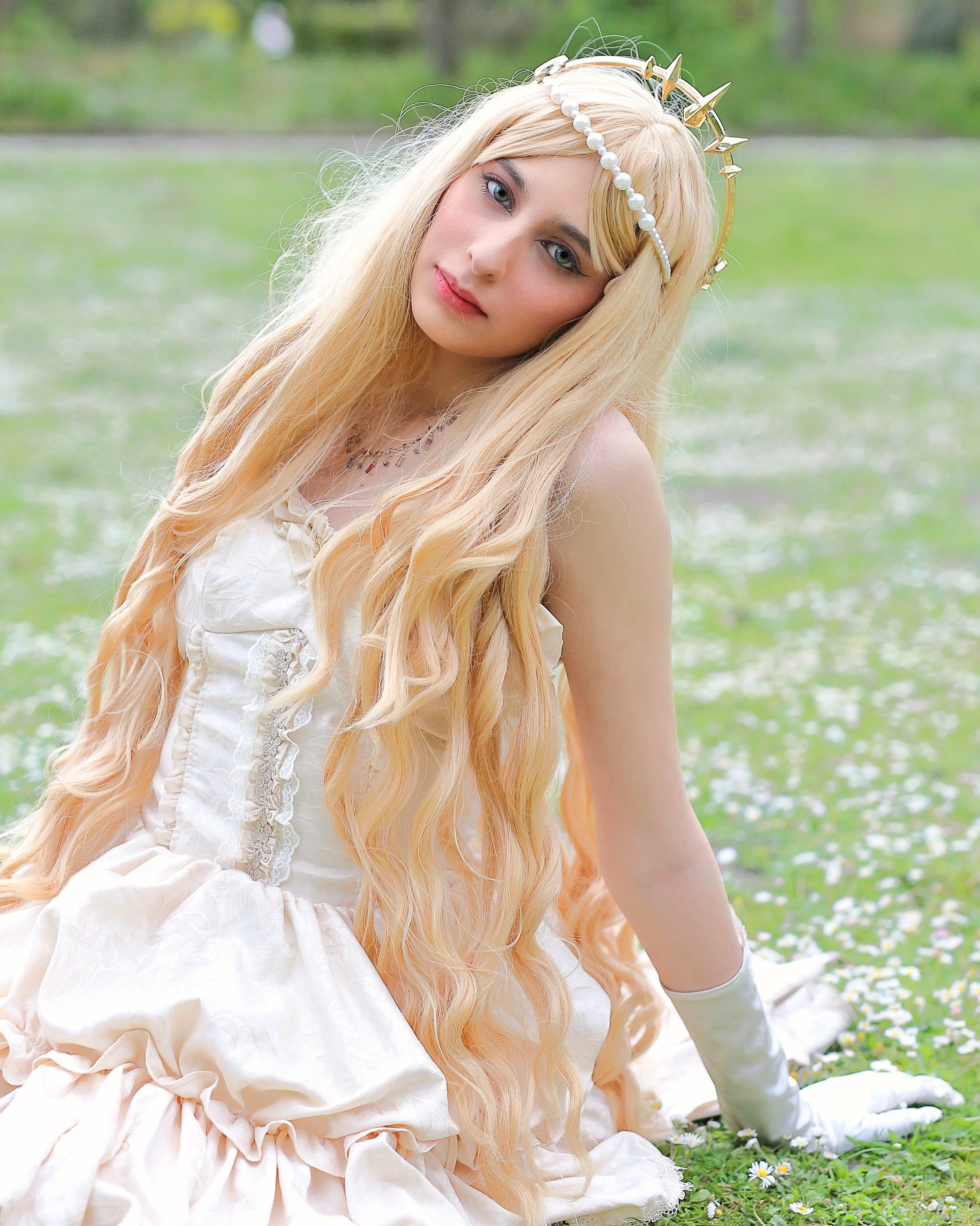 A young woman with long, wavy blonde hair wearing a white dress and a pearl and gold crown, sitting on a grassy field with small white flowers, looking at the camera. cosplay cosplayer anime manga musical aphrodite