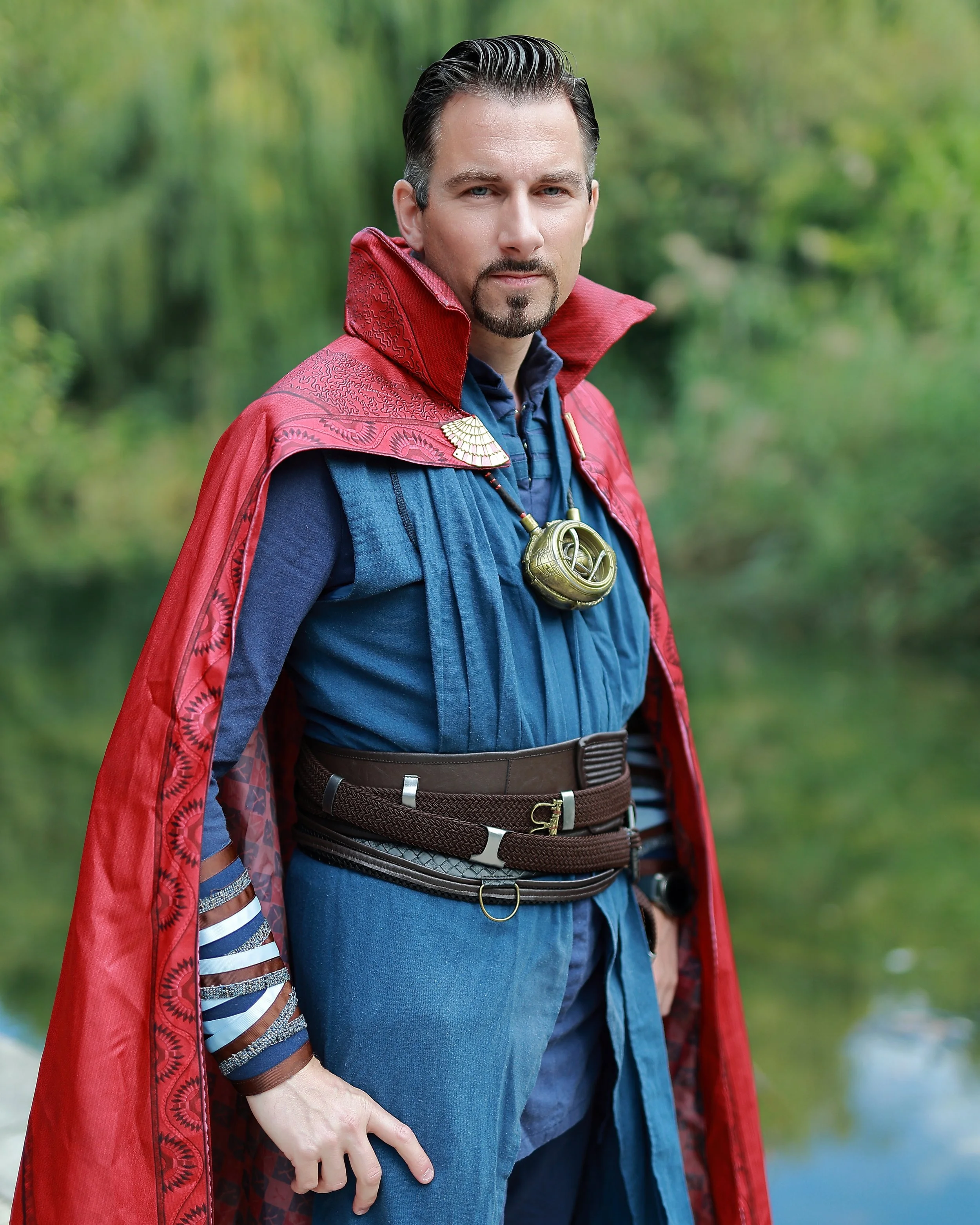 A man dressed as Doctor Strange wearing a red and blue costume with a cape, standing outdoors near a body of water. Cosplay superhero