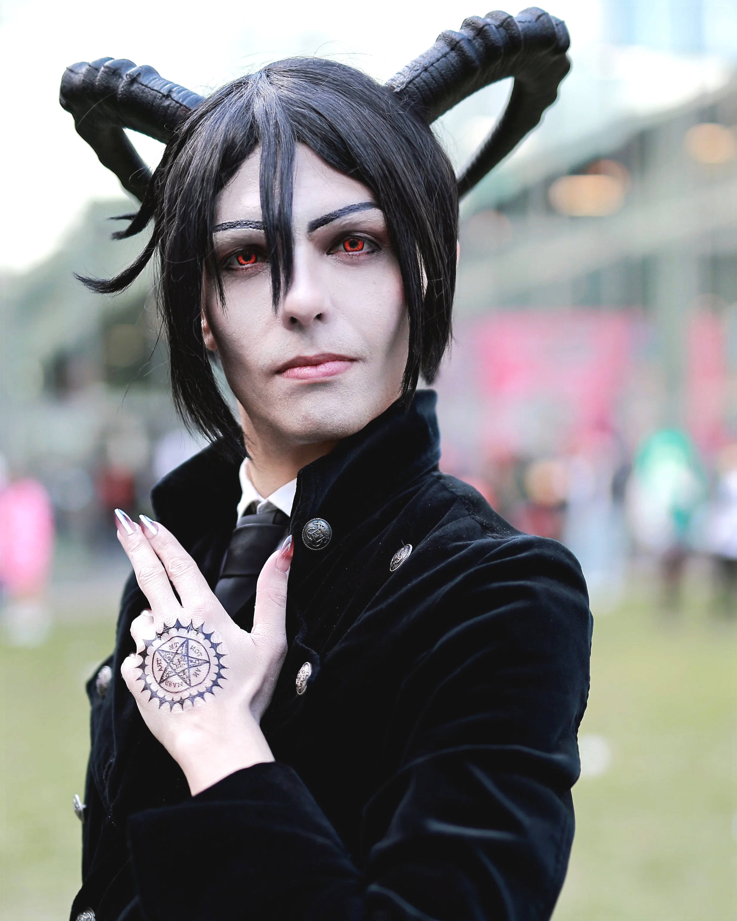 Person with black hair, red eyes, wearing black horns, black coat, showing a pentagram tattoo on hand, outdoors in daytime. cosplay cosplayer manga anime black butler Sebastian