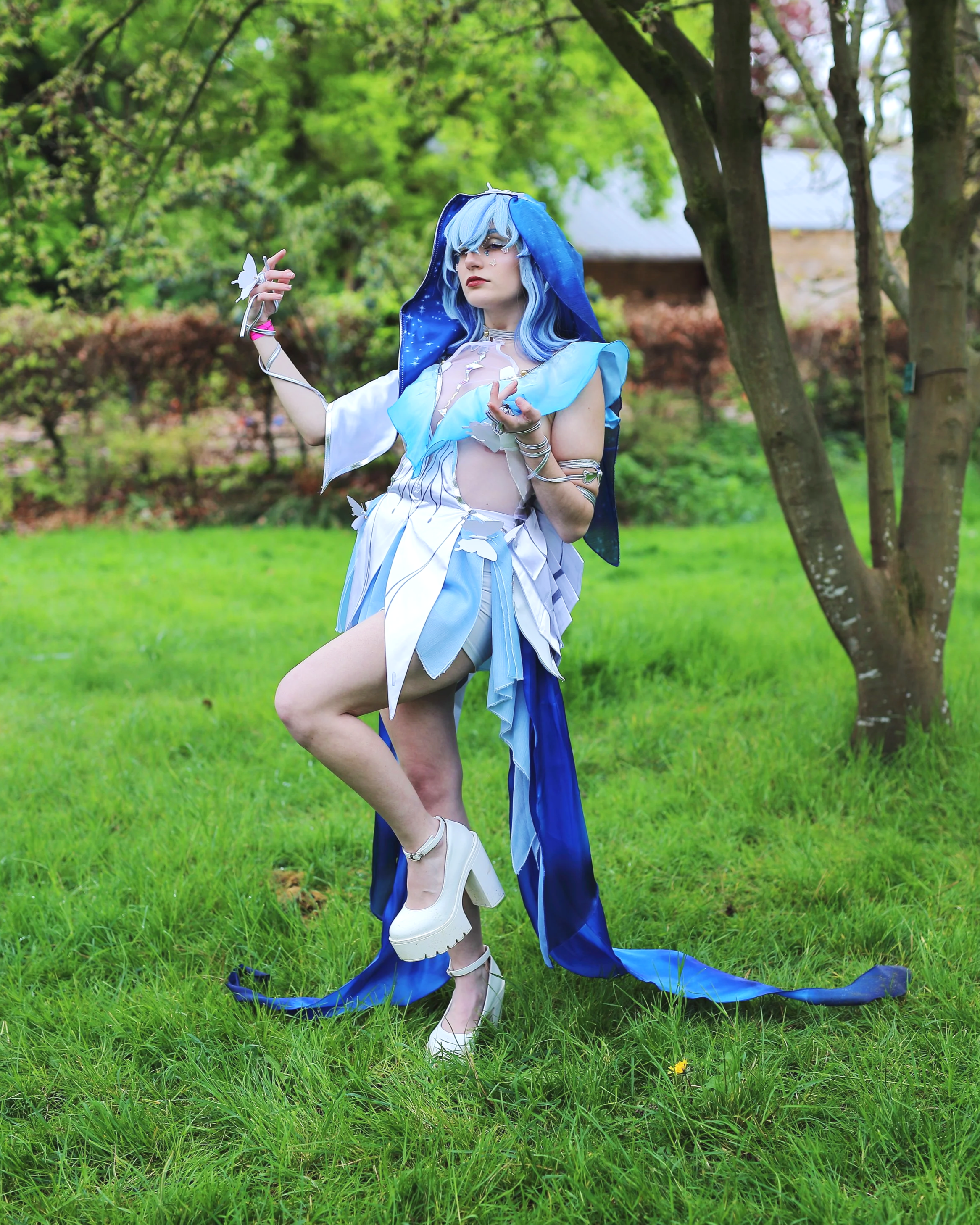 A person dressed in a fantasy or cosplay costume with blue hair, standing on grass in a park with trees in the background. cosplayer anime gaming manga