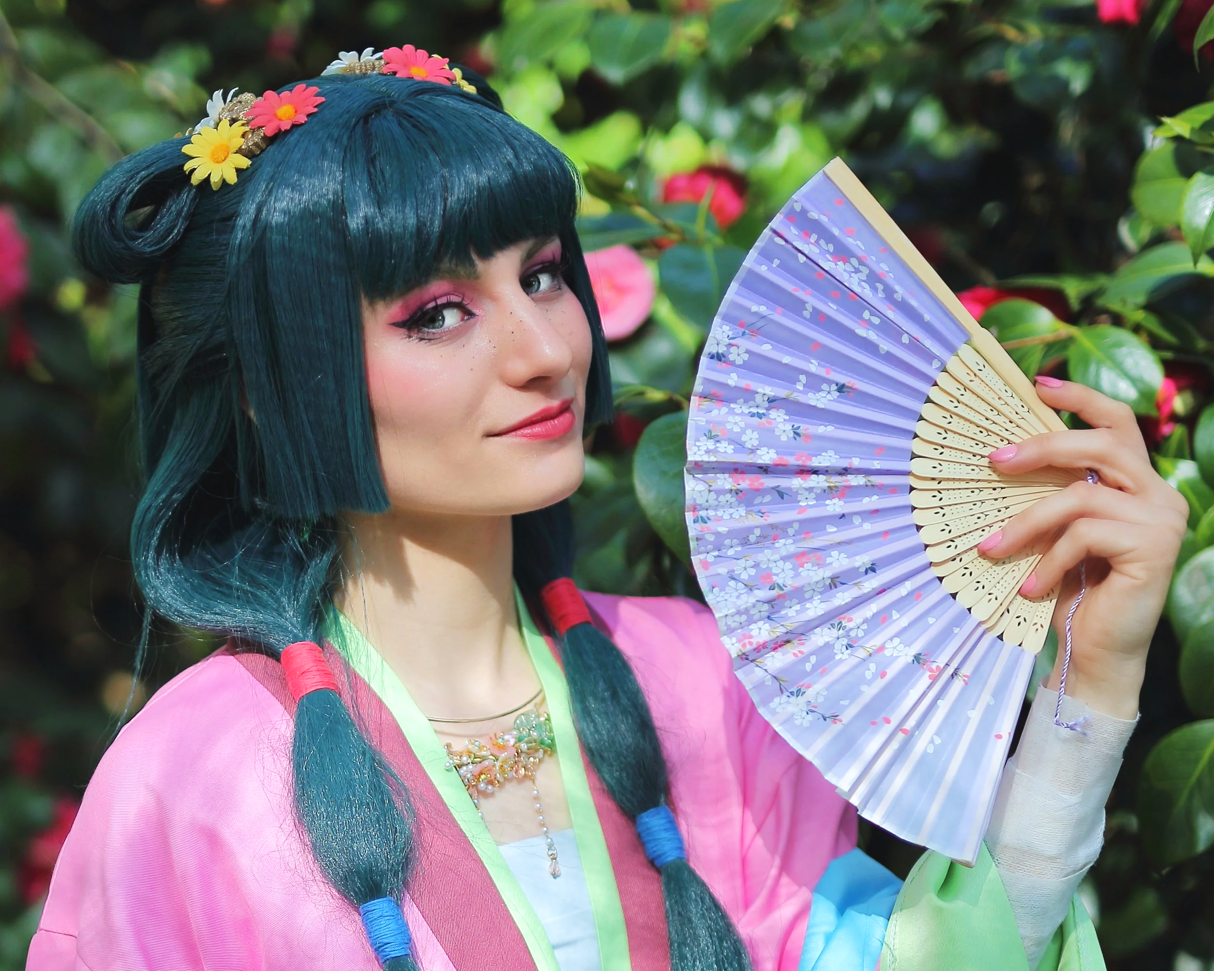 Young woman with dark teal hair styled in two braided pigtails, wearing pink makeup and a colorful flower hairband, holding a purple floral folding fan, standing in front of green foliage with pink flowers. MaoMao, carnets de l'hapothicaire Cosplay