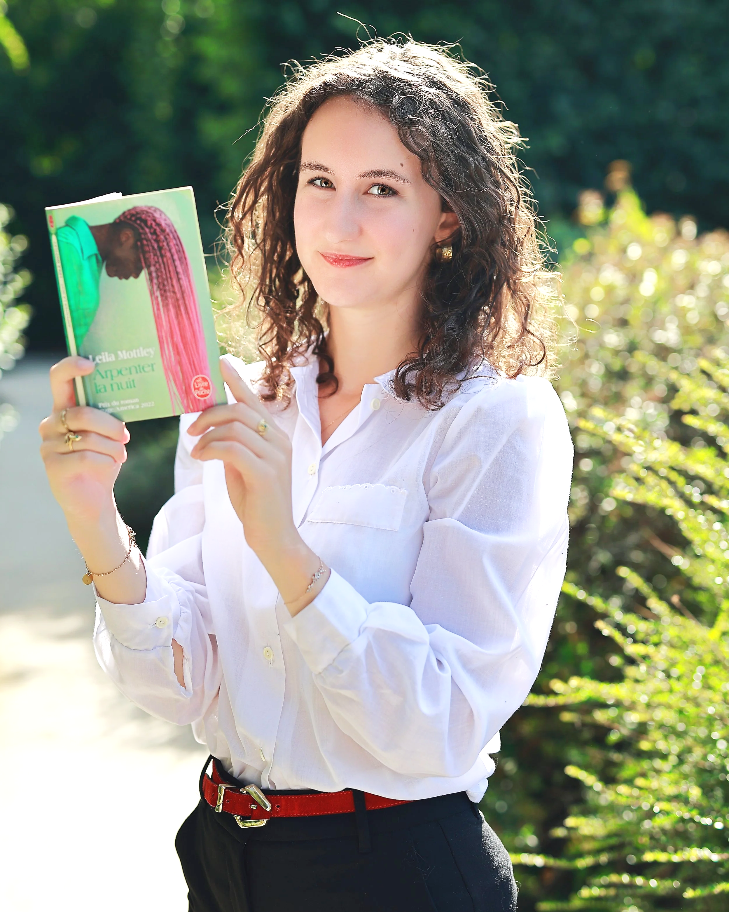 A woman with curly brown hair, wearing a white blouse and black pants, stands outdoors holding a book with a green cover that has a woman with braided hair on it. The background is blurred greenery and sunlight. Portrait