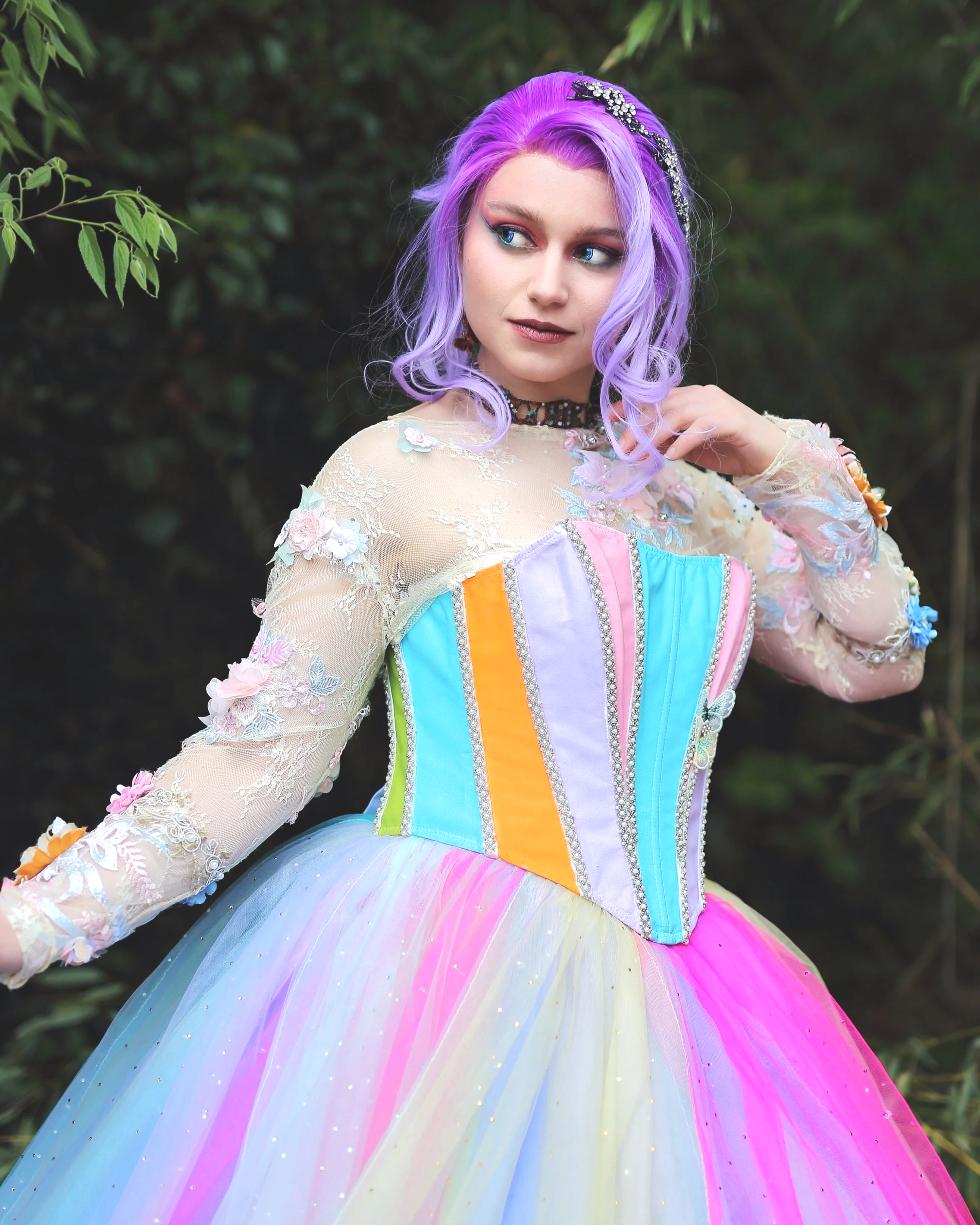 Une femme aux cheveux violets, vêtue d'une robe aux couleurs pastel de l'arc-en-ciel ornée de fleurs et de dentelle, se tient à l'extérieur, devant un parterre de verdure cosplay cosplayer cosplayeuse animation
