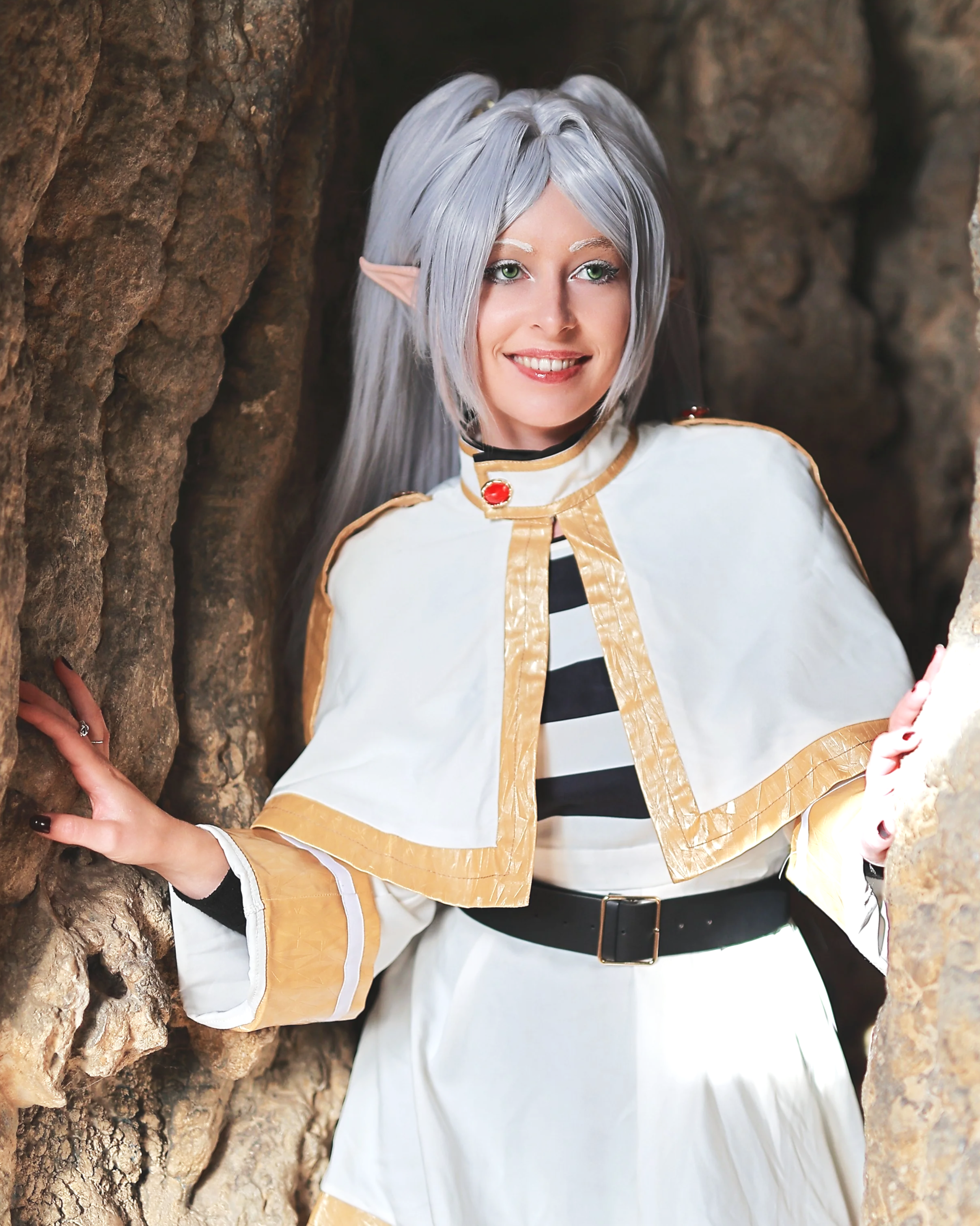 Une femme déguisée en elfe, en Frieren ou en personnage fantastique, avec des cheveux gris clair, des oreilles d'elfe et un costume blanc et or, se tient dans un décor rocheux. Cosplayeuse Anime Manga cosplay cosplayer déguisement