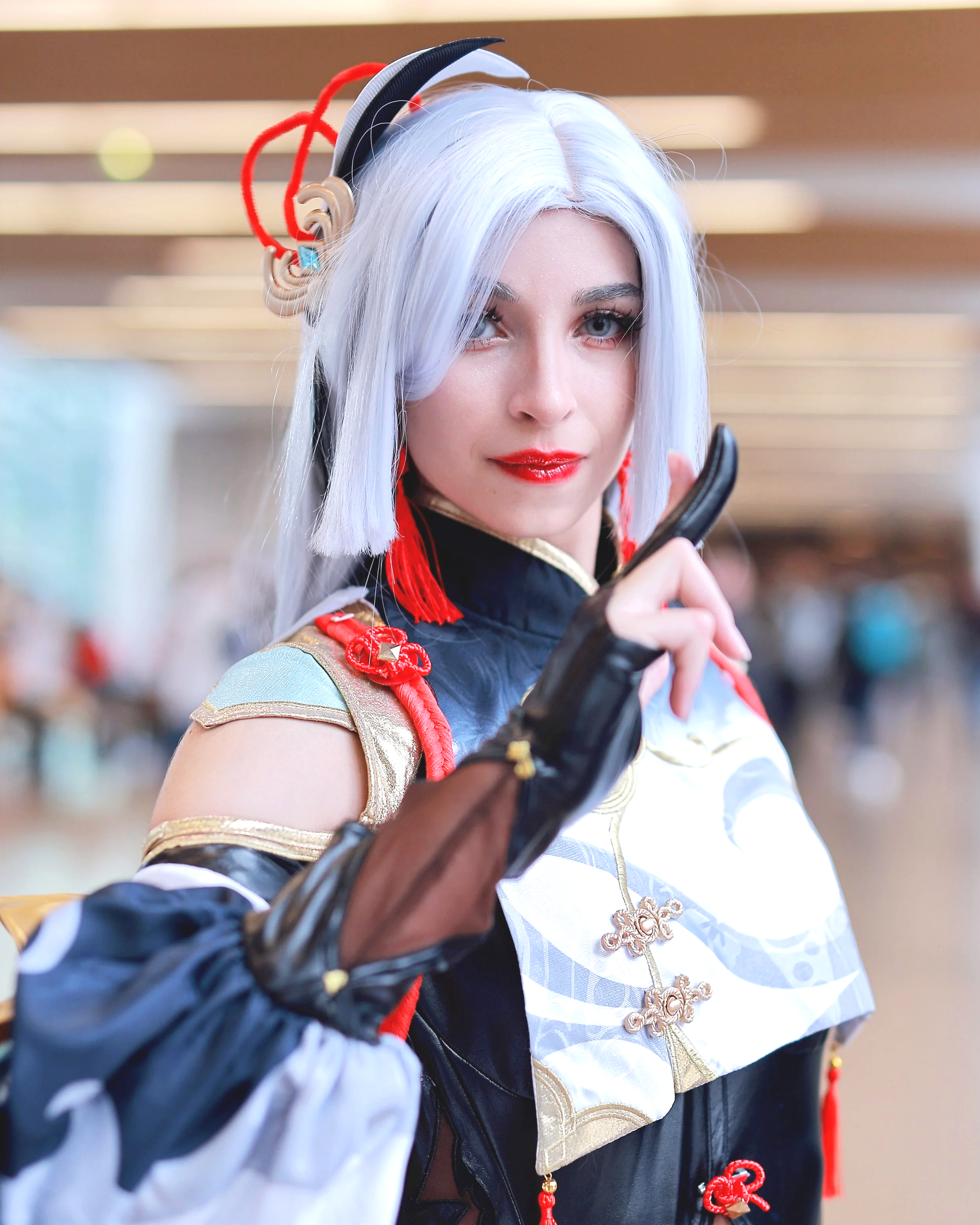 A person dressed in a detailed costume with white hair, red accents, and gold embellishments, holding a black prop and looking into the camera. Cosplay anime manga gaming
