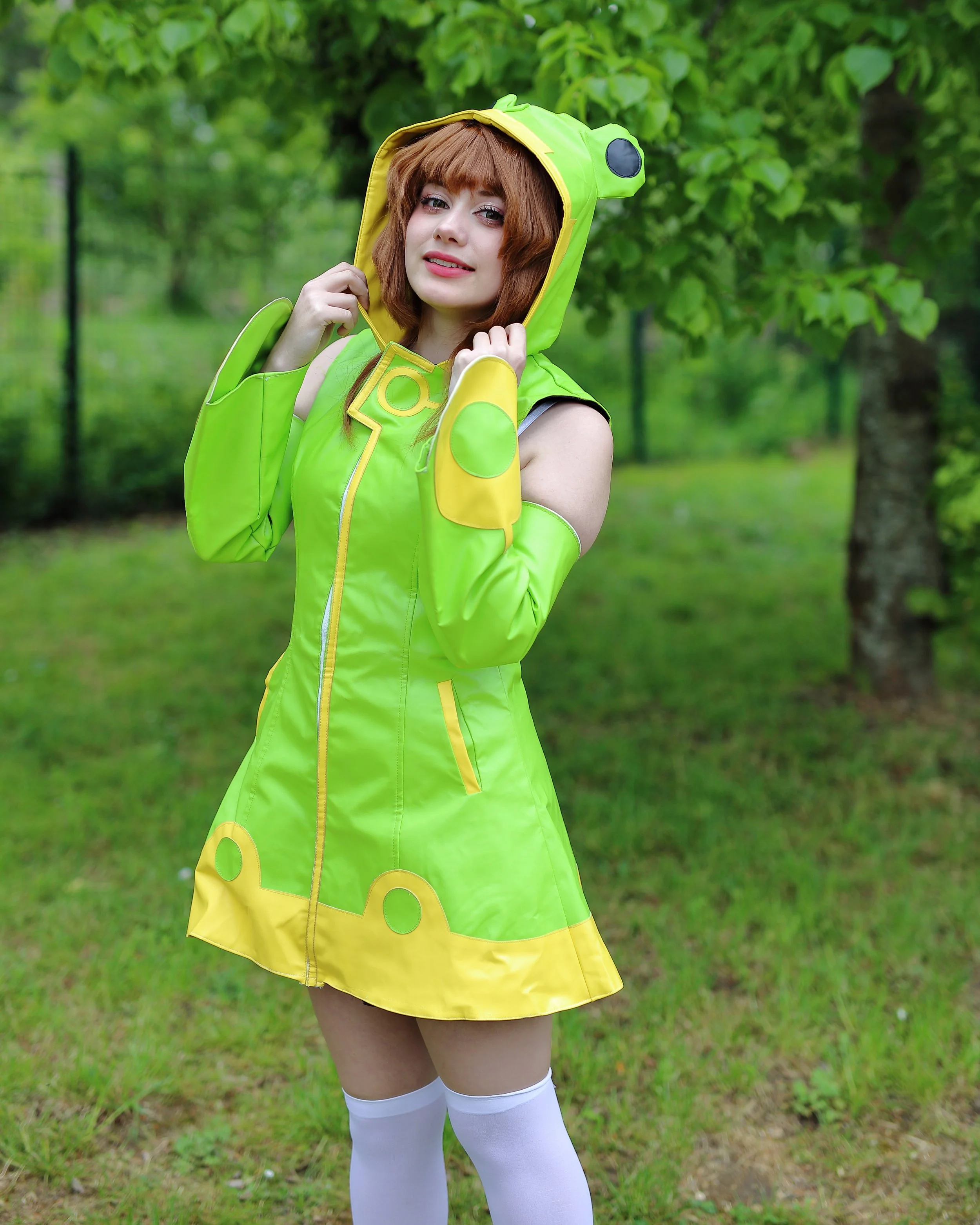 A young woman with auburn hair in a green and yellow raincoat with a bear design, standing outdoors in a park with green trees, holding the hood of her raincoat. cosplay cosplayer manga anime gaming