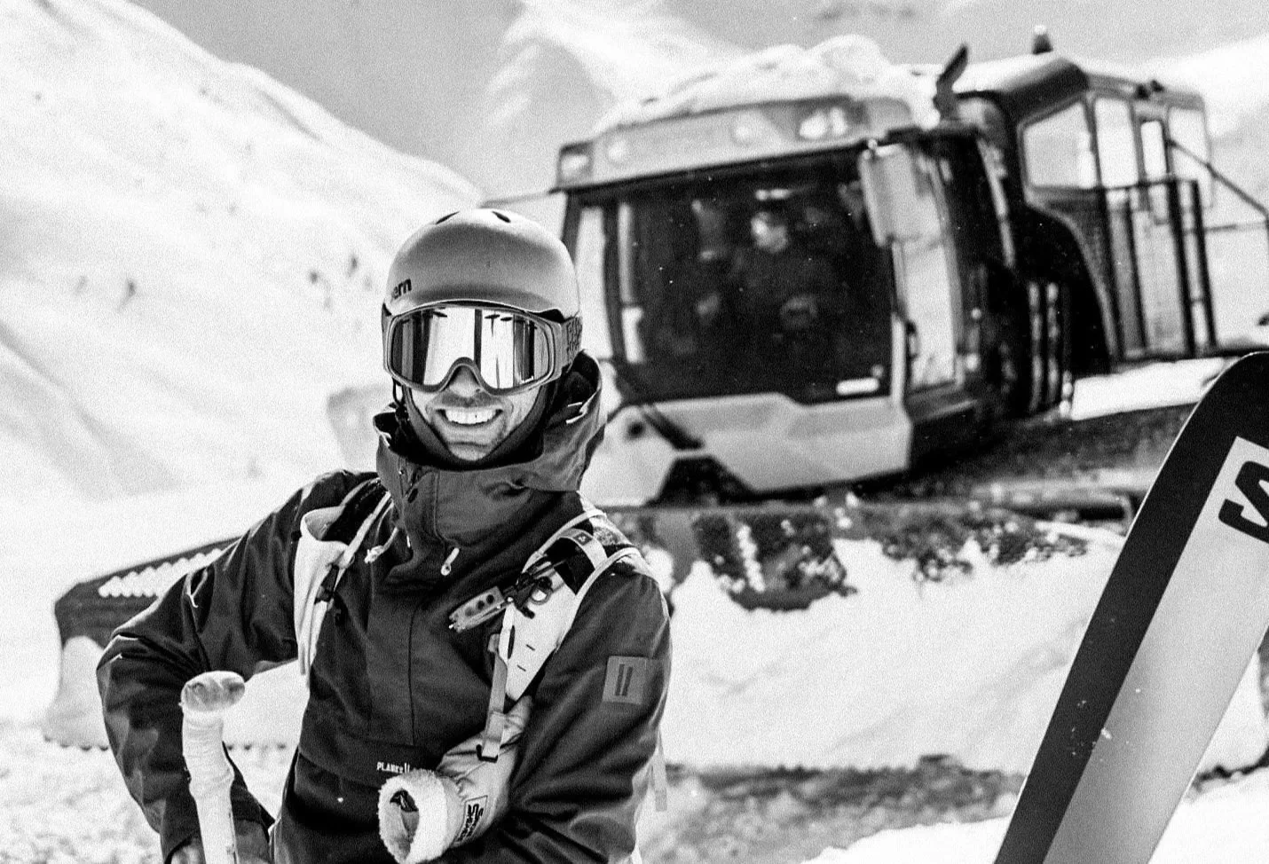 A man outdoors in a snowy environment, dressed in winter gear, including a dark jacket with patches, a helmet, sunglasses, gloves, and holding ski poles, smiling.
