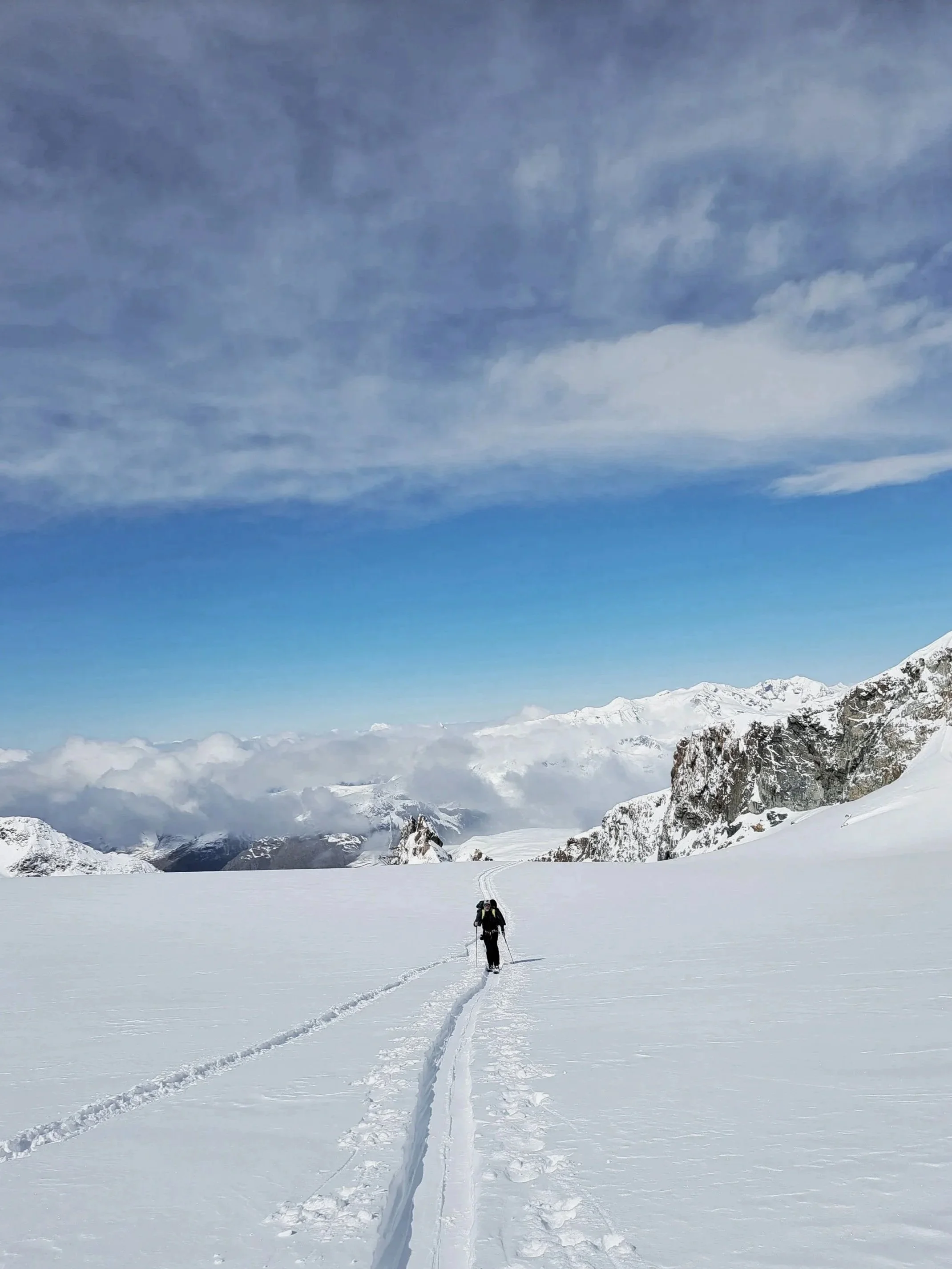 A person cross-country skiing in a vast, snowy mountain landscape with cloudy skies.
