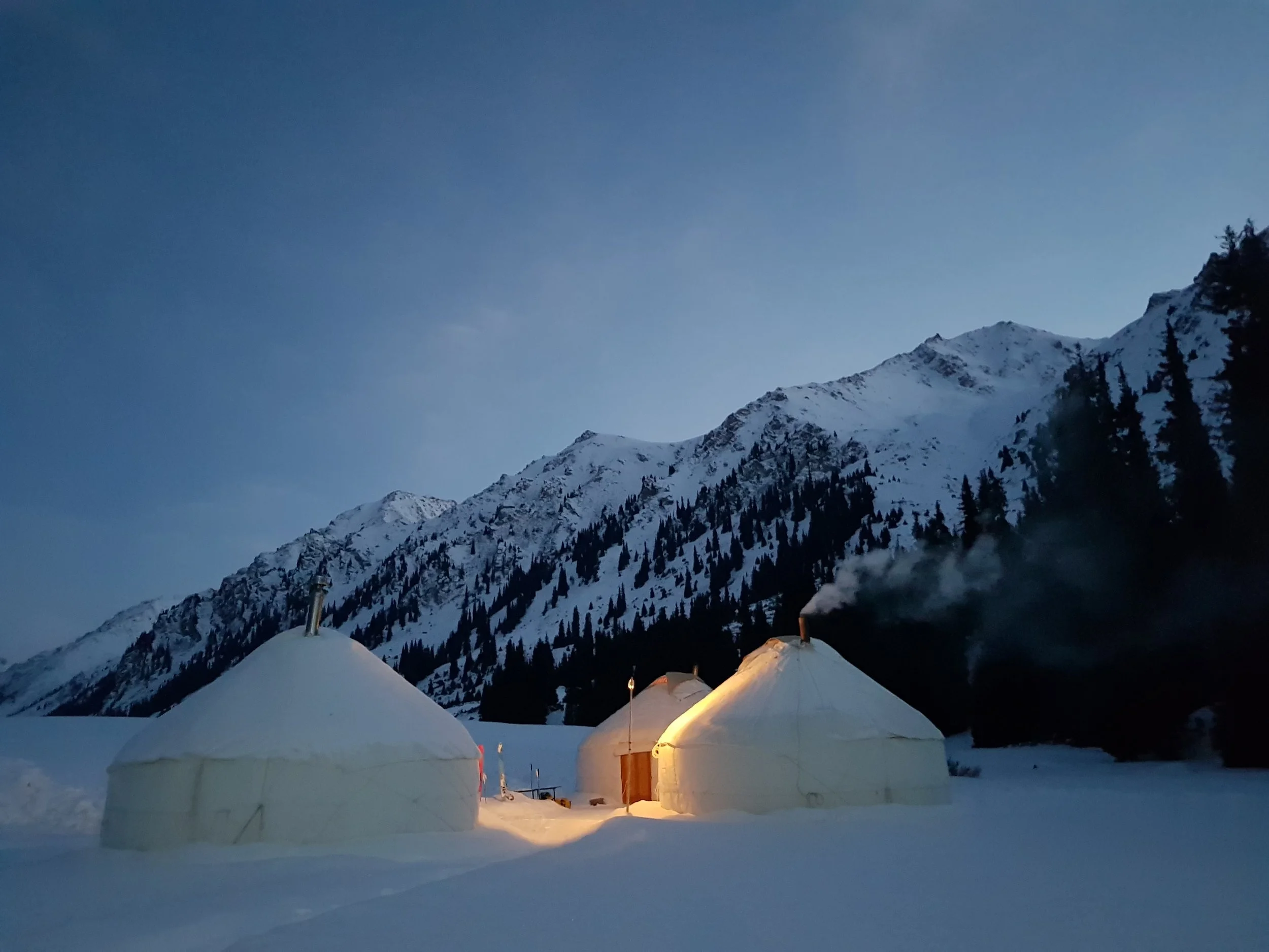 Snow-covered tents in a mountainous landscape with pine trees and a clear twilight sky, smoke rising from one of the tents.