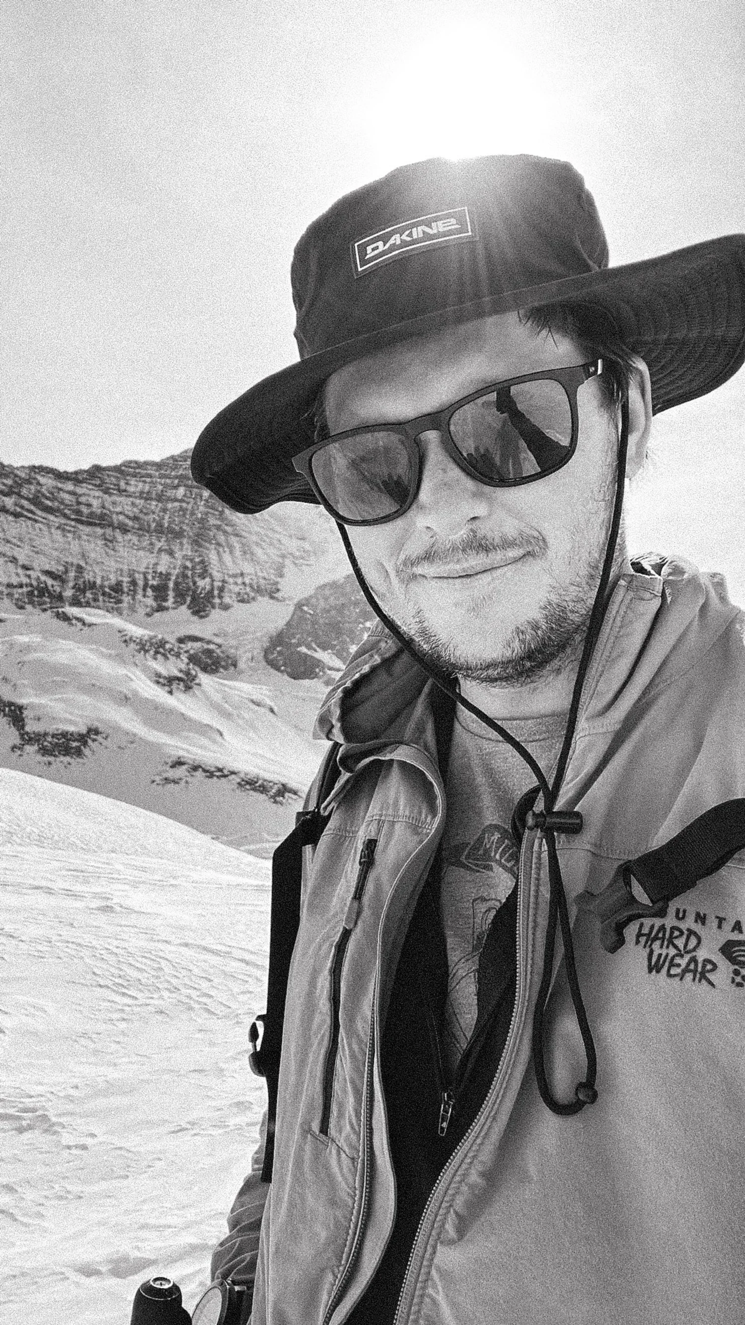 A man in winter gear and sunglasses standing on snow with snowy mountain landscape in the background.