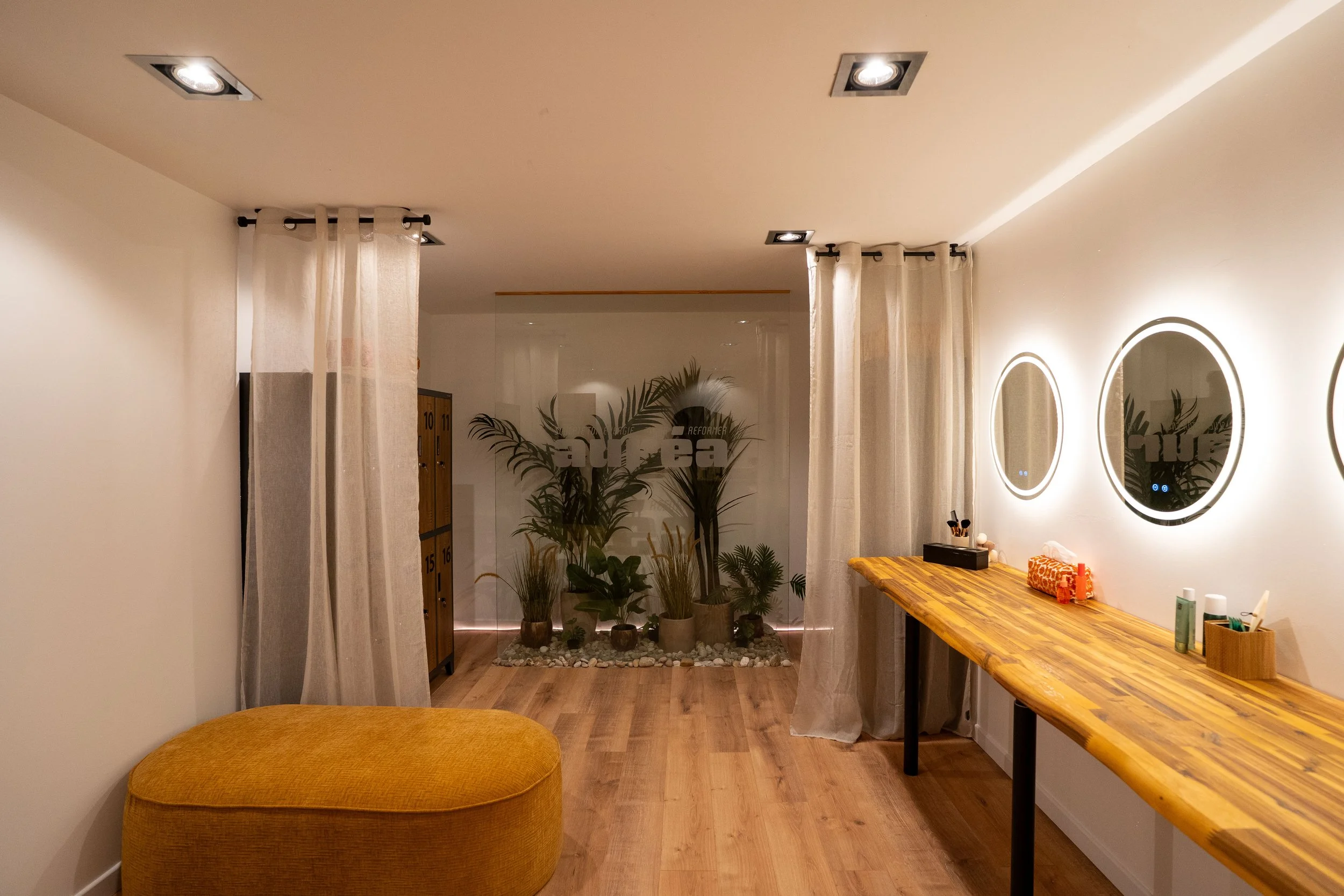 Vestiaire femmes du studio Auréa, un cadre chaleureux avec des mirroir eclairé de la végétation derriere une vitre dans le fond et des fauteuils jaune pour s'asseoir. Casier en simili bois