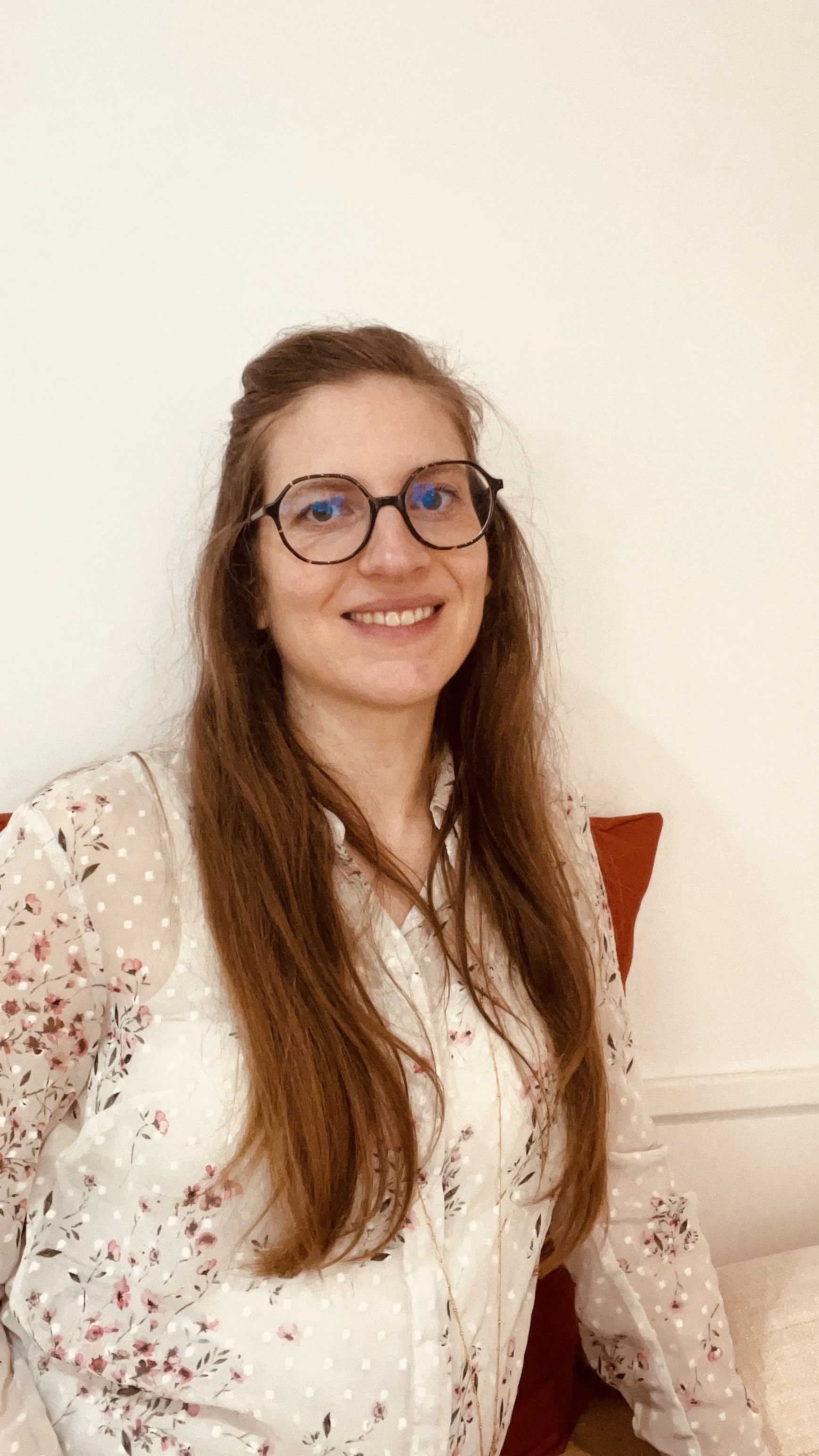 Jeune femme aux longs cheveux roux, portant des lunettes rondes, souriante, portant une chemise blanche à motifs floraux, assise sur un canapé avec un coussin rouge, contre un mur blanc.