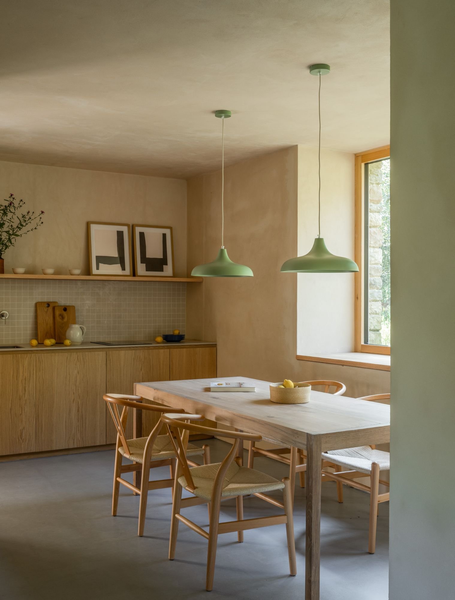 View of a minimalist kitchen with light wood cabinets, decorative objects and plants on a shelf, and a wooden table with wooden chairs next to a beige wall.
