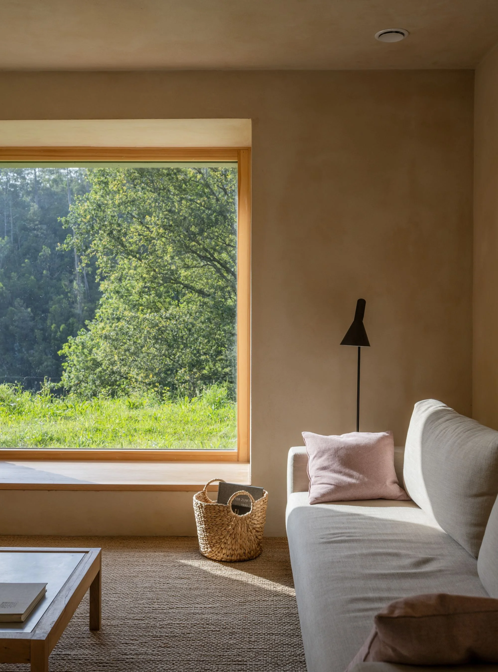 Room with large window overlooking green trees, beige sofa with pink cushion, black floor lamp, wicker basket with books, fiber rug and wooden coffee table.
