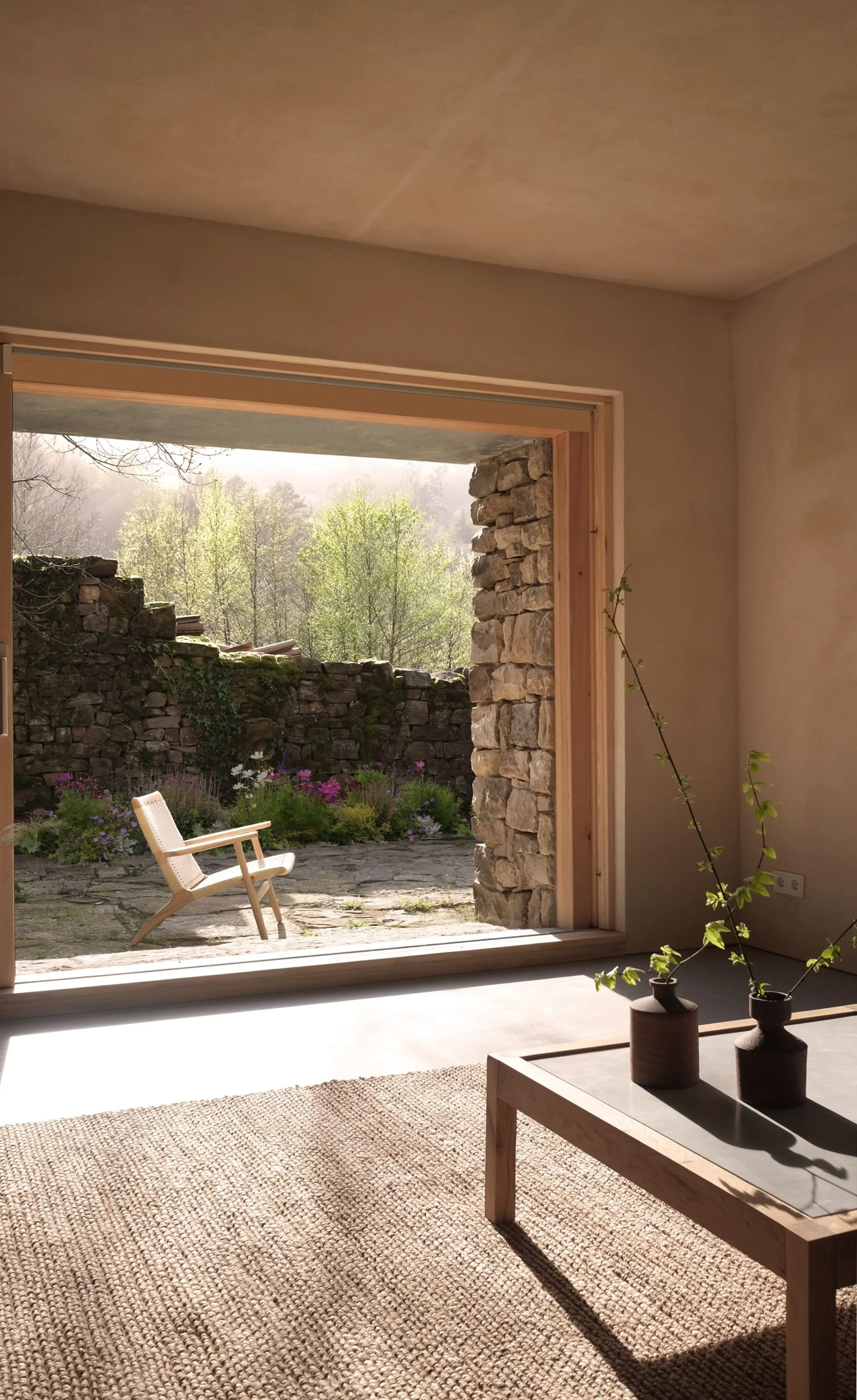 Vista desde el interior de una casa con paredes de color beige, una mesa de madera con dos jarrones con plantas y una ventana grande que muestra un jardín con flores, un muro de piedra y árboles verdes en el fondo.