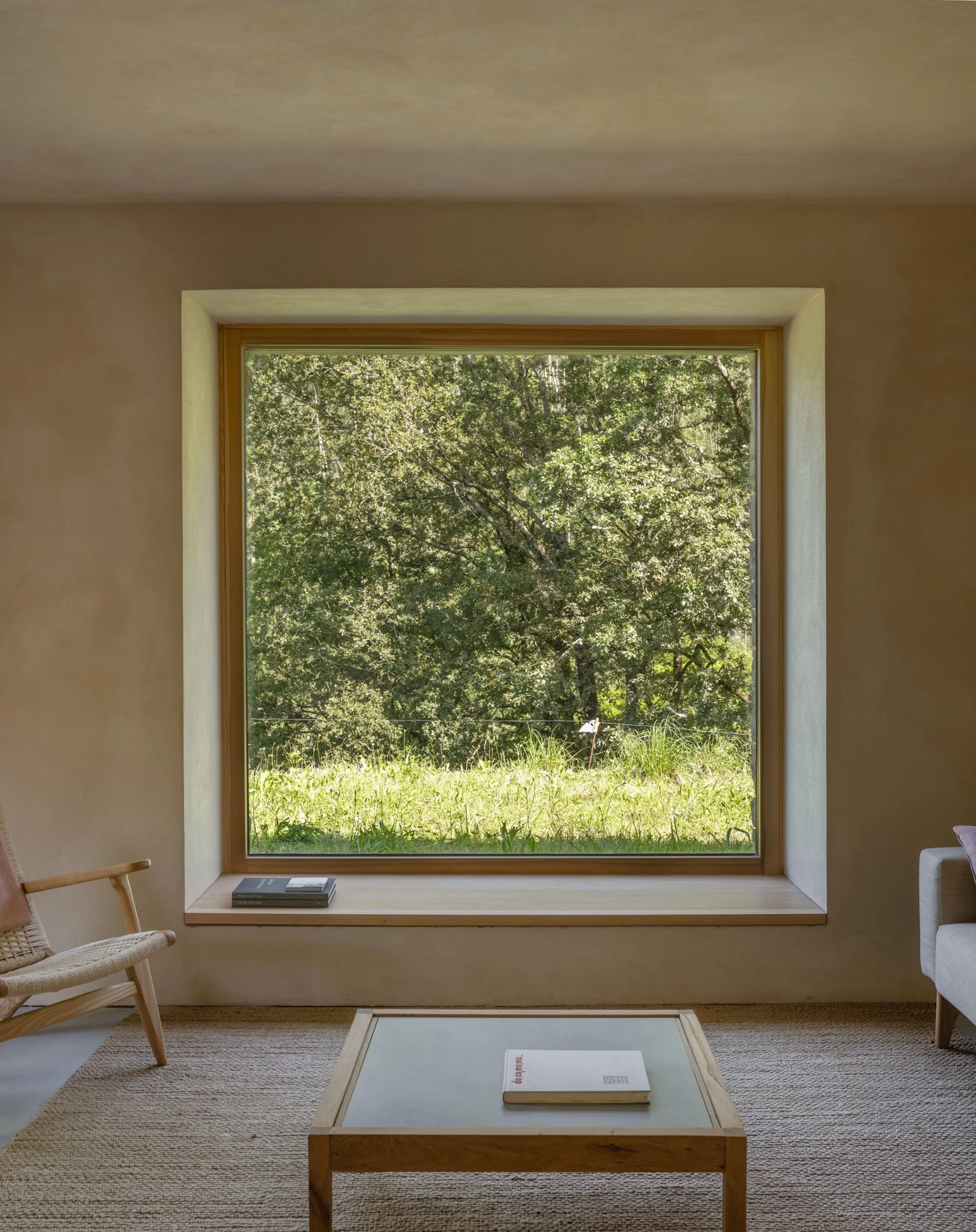 Room with large window overlooking green trees, beige sofa with pink cushion, black floor lamp, wicker basket with books, fiber rug and wooden coffee table.