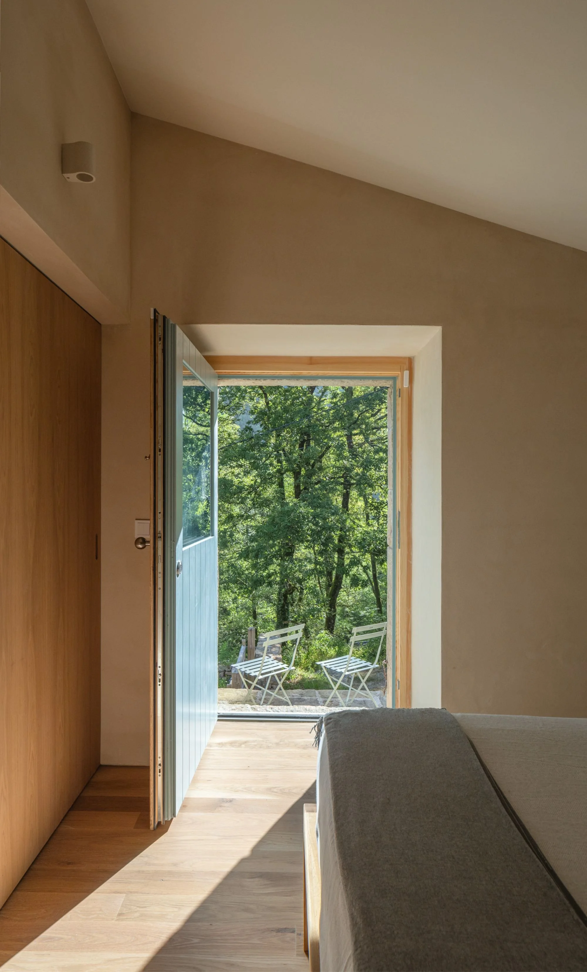 View from a room with an open door leading to a terrace with white chairs and green trees.