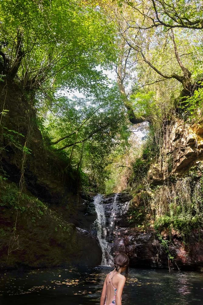 Niña en traje de baño en un río rodeada de vegetación y una pequeña cascada natural.